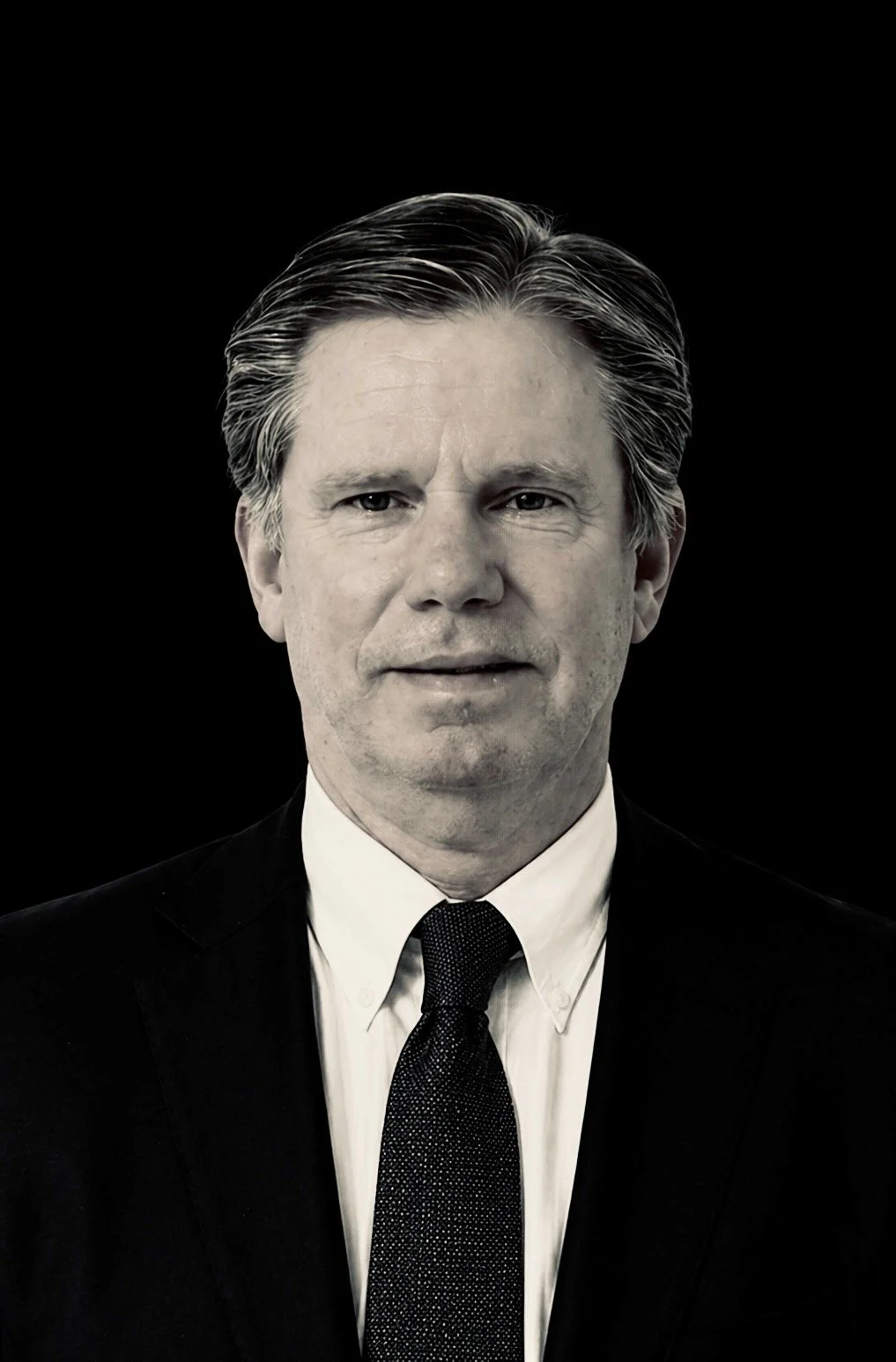 Black and white portrait of a middle-aged man with dark hair, wearing a suit and tie, against a black background.