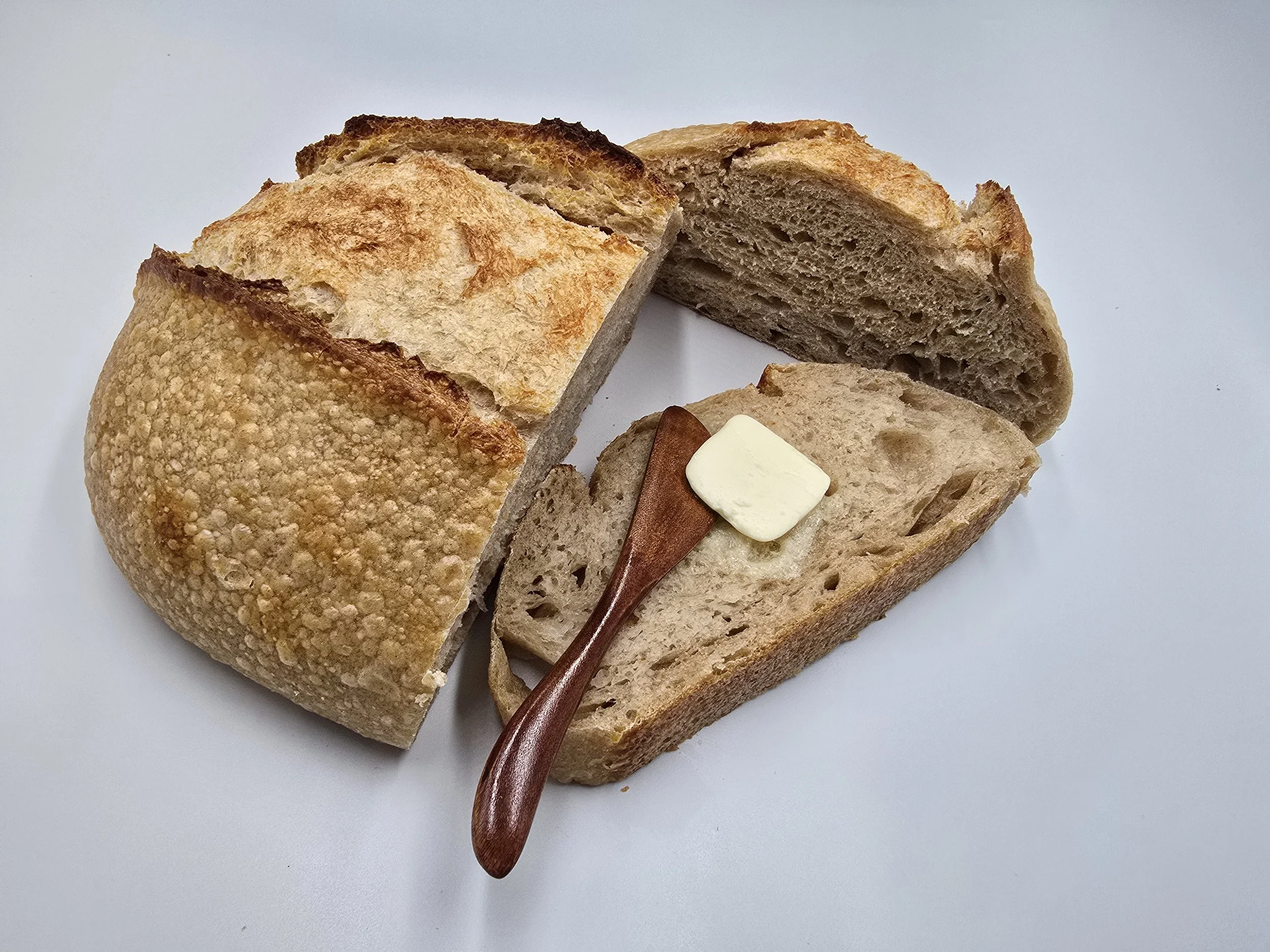 A loaf of rustic bread with a crispy crust, one slice cut and spread with butter, and a small butter knife resting on the slice, all on a white background.