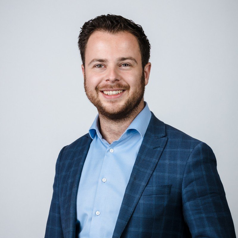 Portret van een lachende man met donker haar, een baard, gekleed in een blauwe blouse en een donkerblauwe geruite jas, tegen een lichte achtergrond. Ruben Paijens uit Rotterdam, eigenaar van COHORT en CADRE.