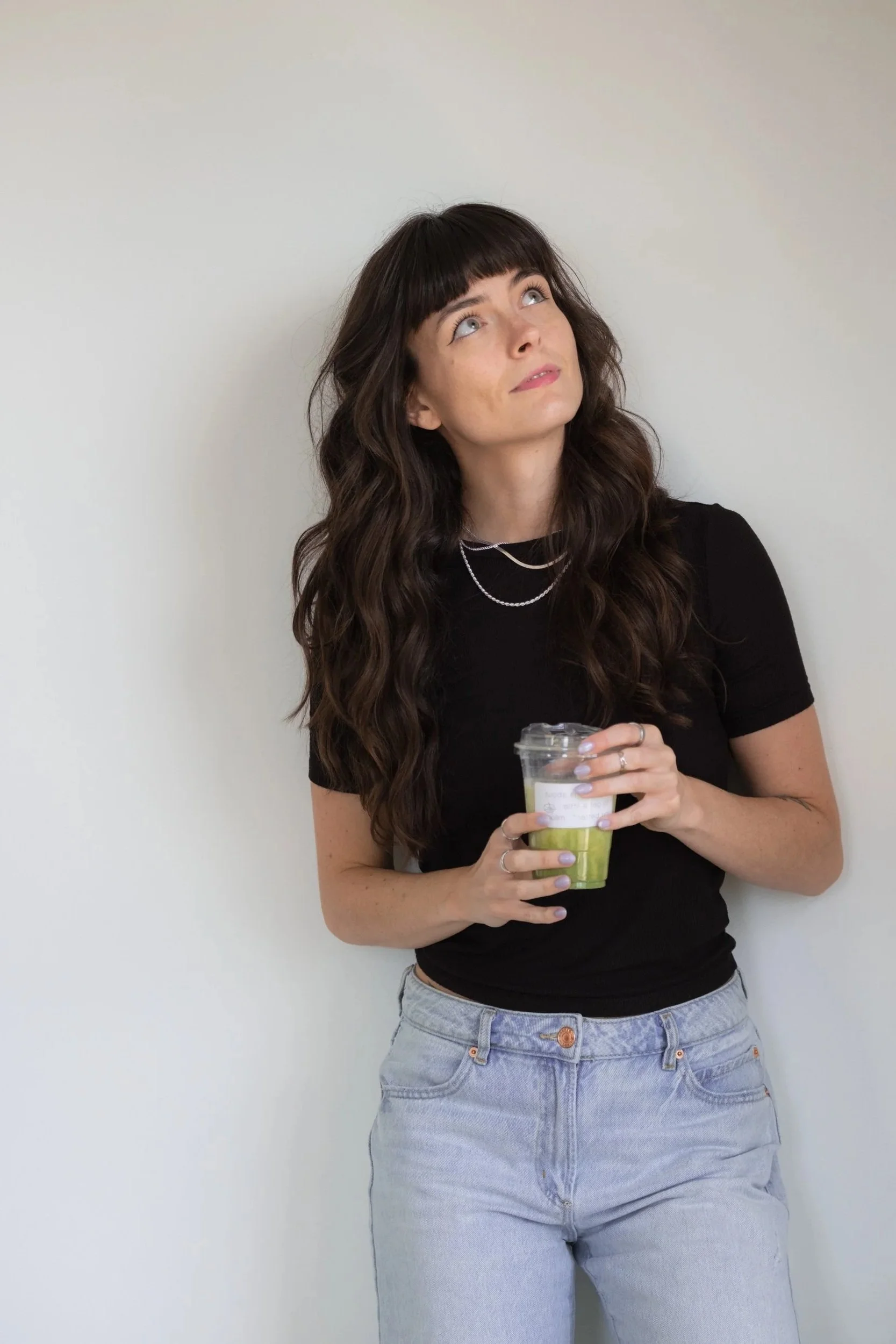 A woman with long wavy dark hair and bangs, wearing a black t-shirt and light blue jeans, holding a plastic cup, standing against a plain white wall, looking up and to her left.