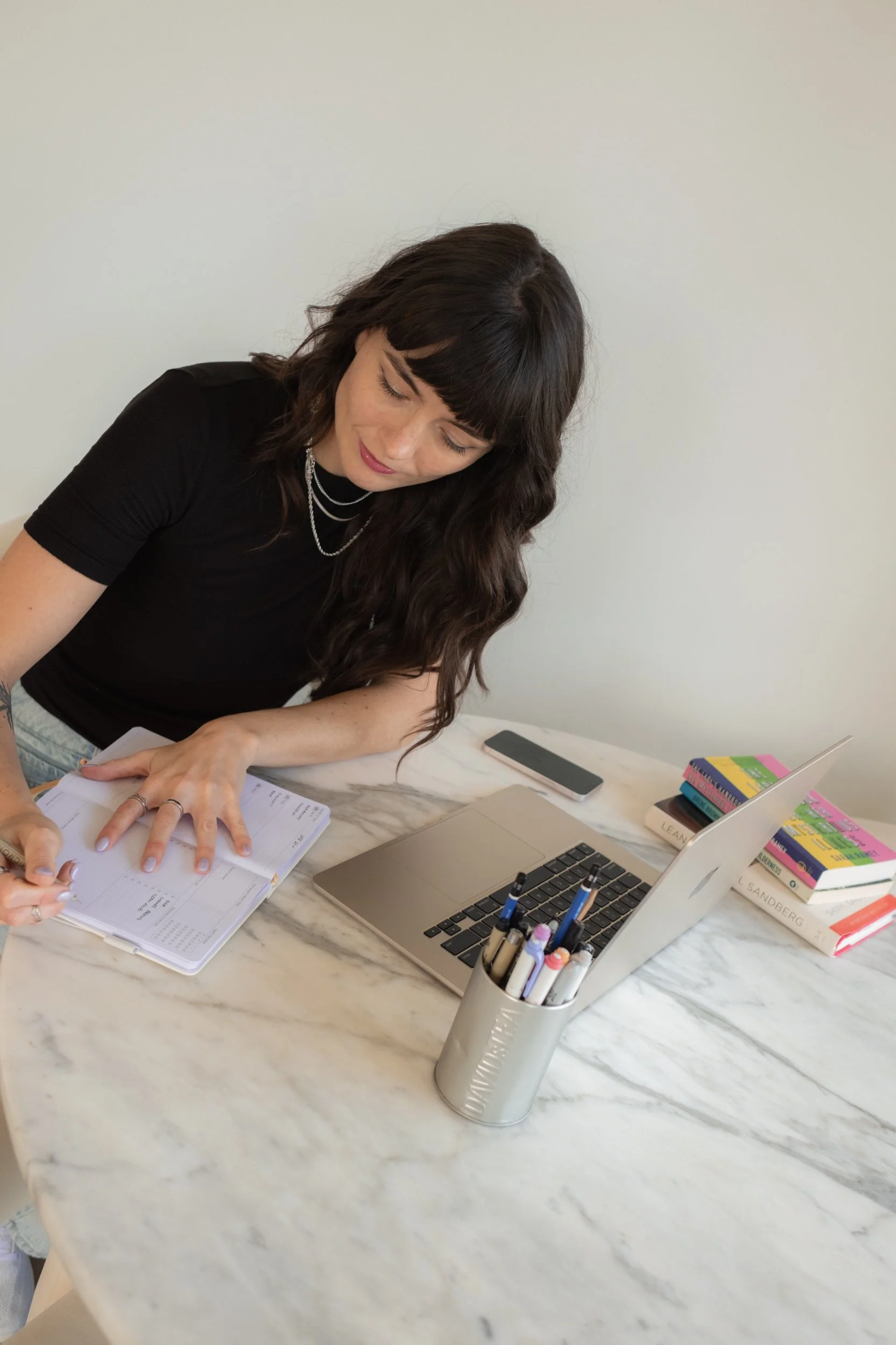 Woman with black hair and a black shirt writing in planner at a white marble desk, with laptop, pens, and books nearby.