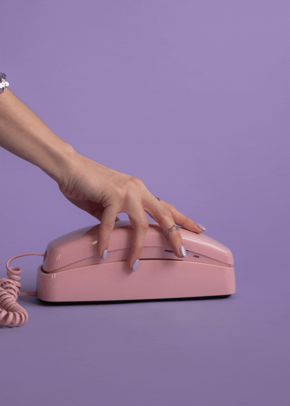 A person's hand with lavender nail polish reaching to press a pink vintage corded telephone on a purple surface with a matching purple background.