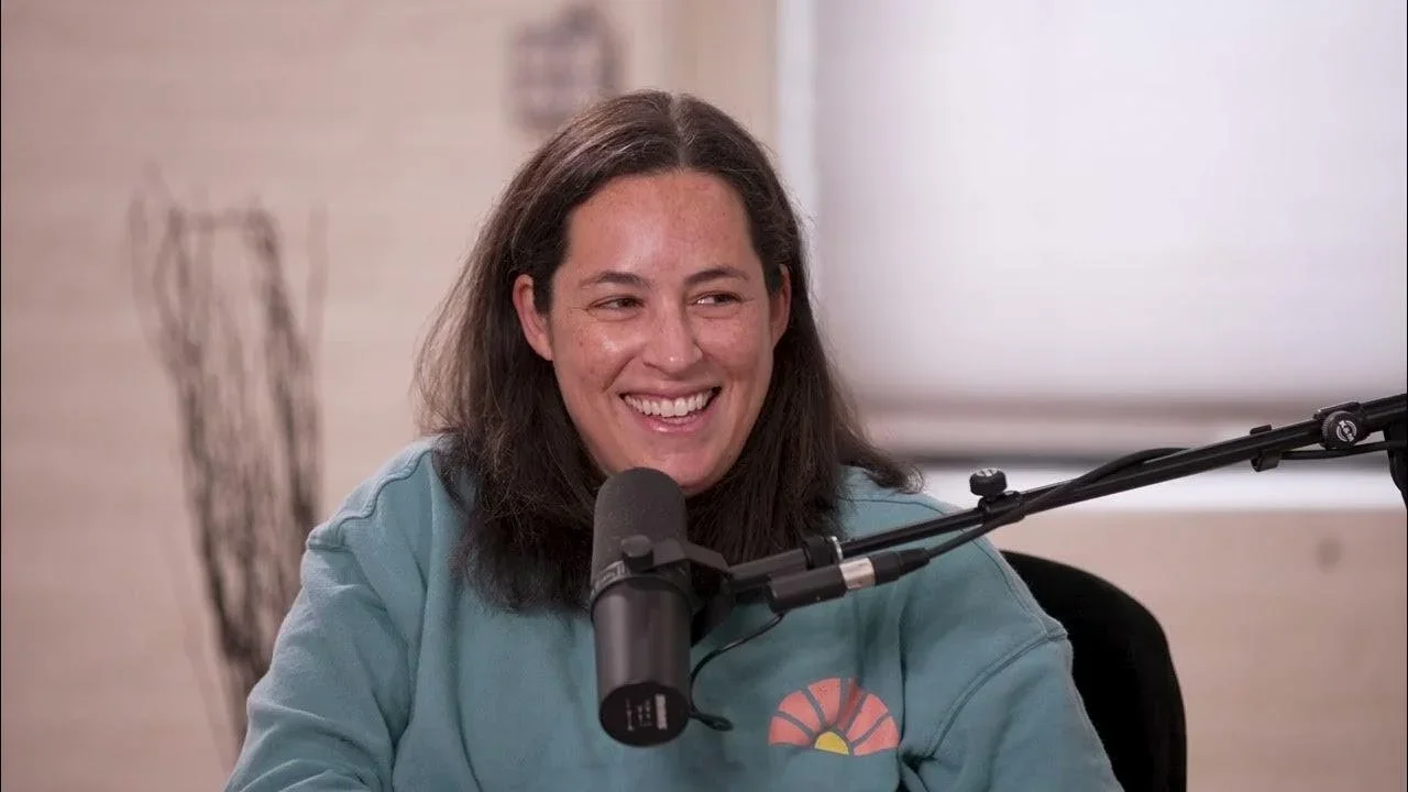 A woman with dark hair smiling, sitting at a table with a microphone in front of her, wearing a blue sweatshirt.