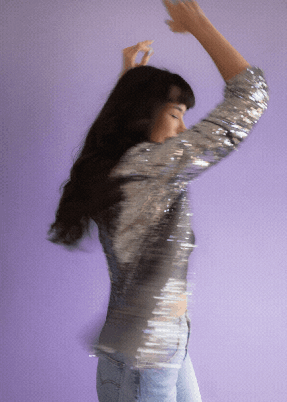 A woman with long dark hair dancing against a purple background, wearing a silver sequined top and light blue jeans.