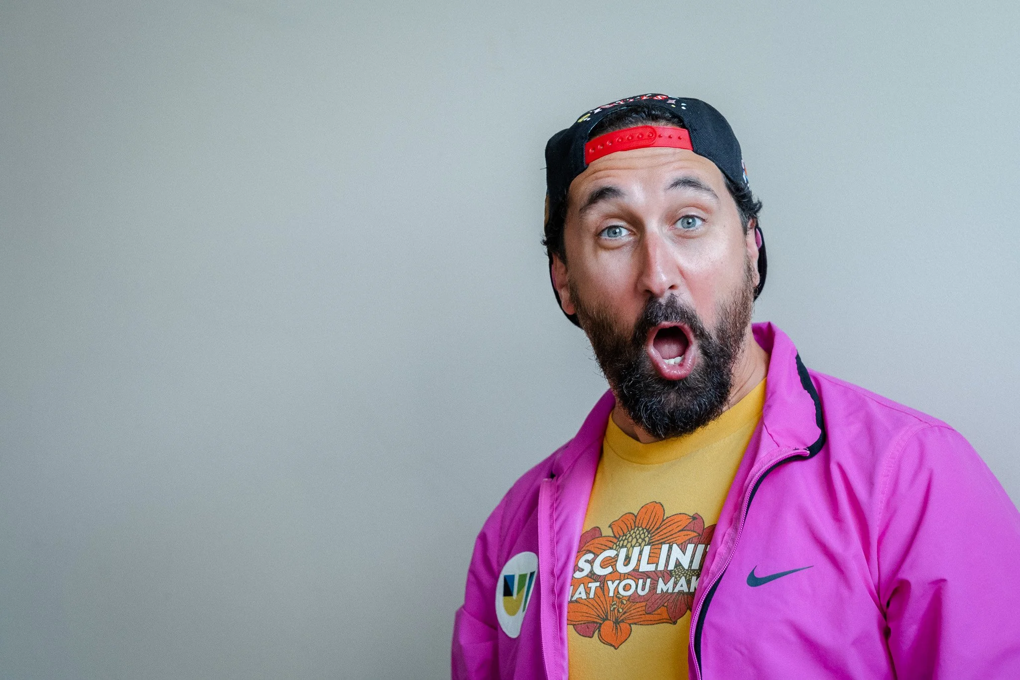 A man with a beard and blue eyes wearing a black and red backwards cap, a purple jacket with Nike logo, and a yellow T-shirt with floral design and the text Masculinity is What You Make", appears surprised and happy.