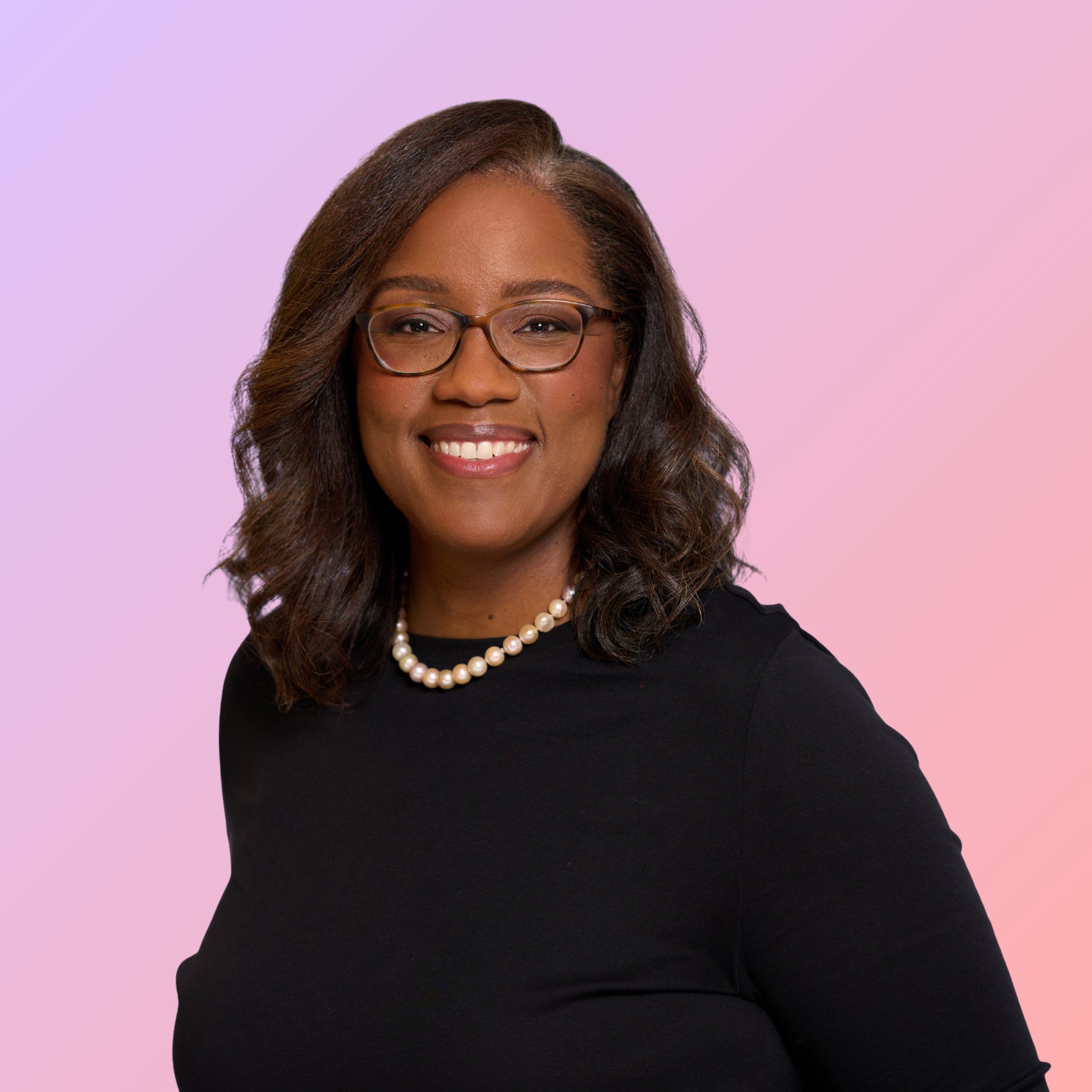 Portrait of a smiling woman with glasses, pearl necklace, and black top against a pastel pink and purple background.