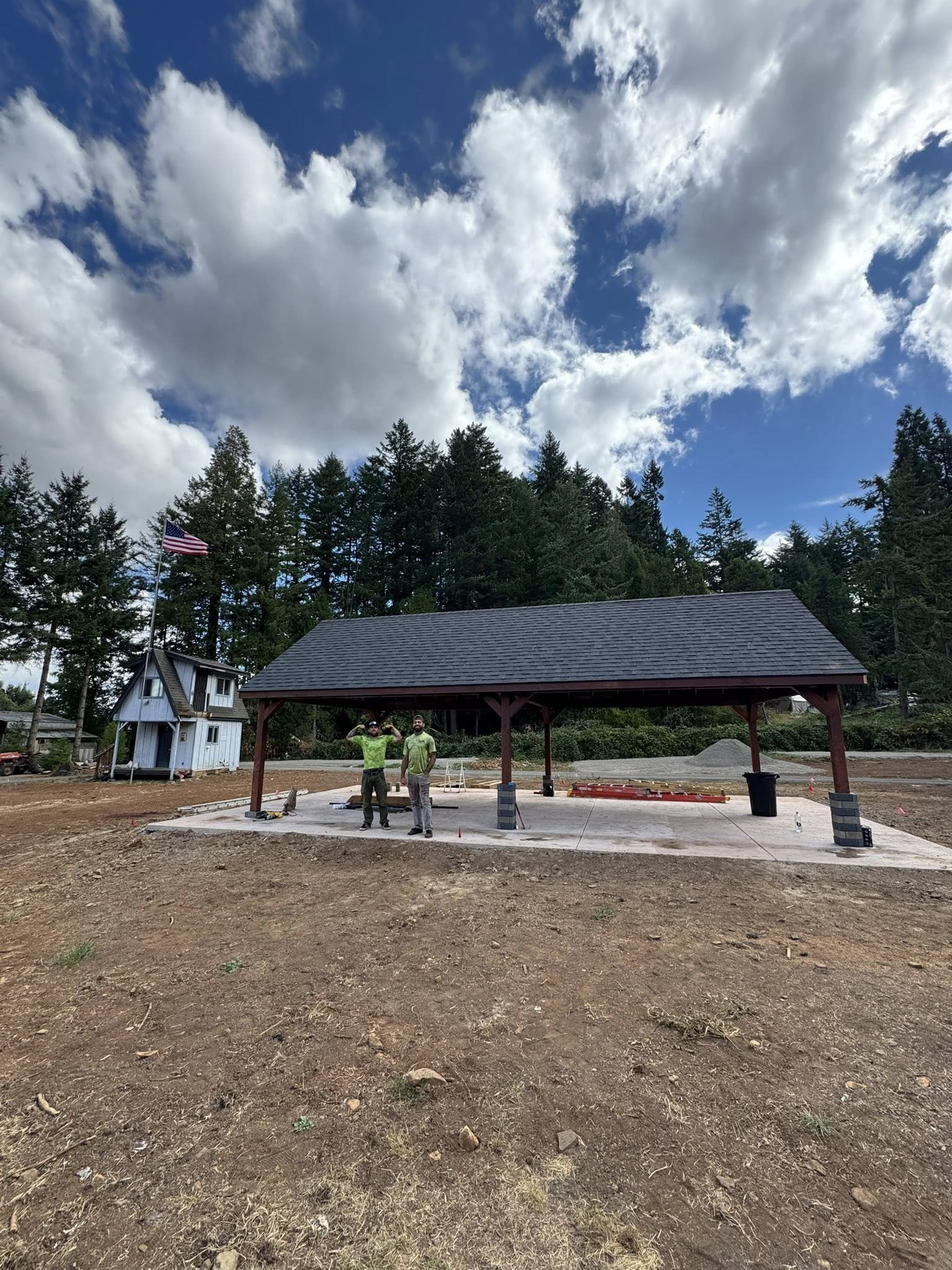Construction workers standing under a small pavilion with a sloped roof, with a wooded background and partly cloudy sky.