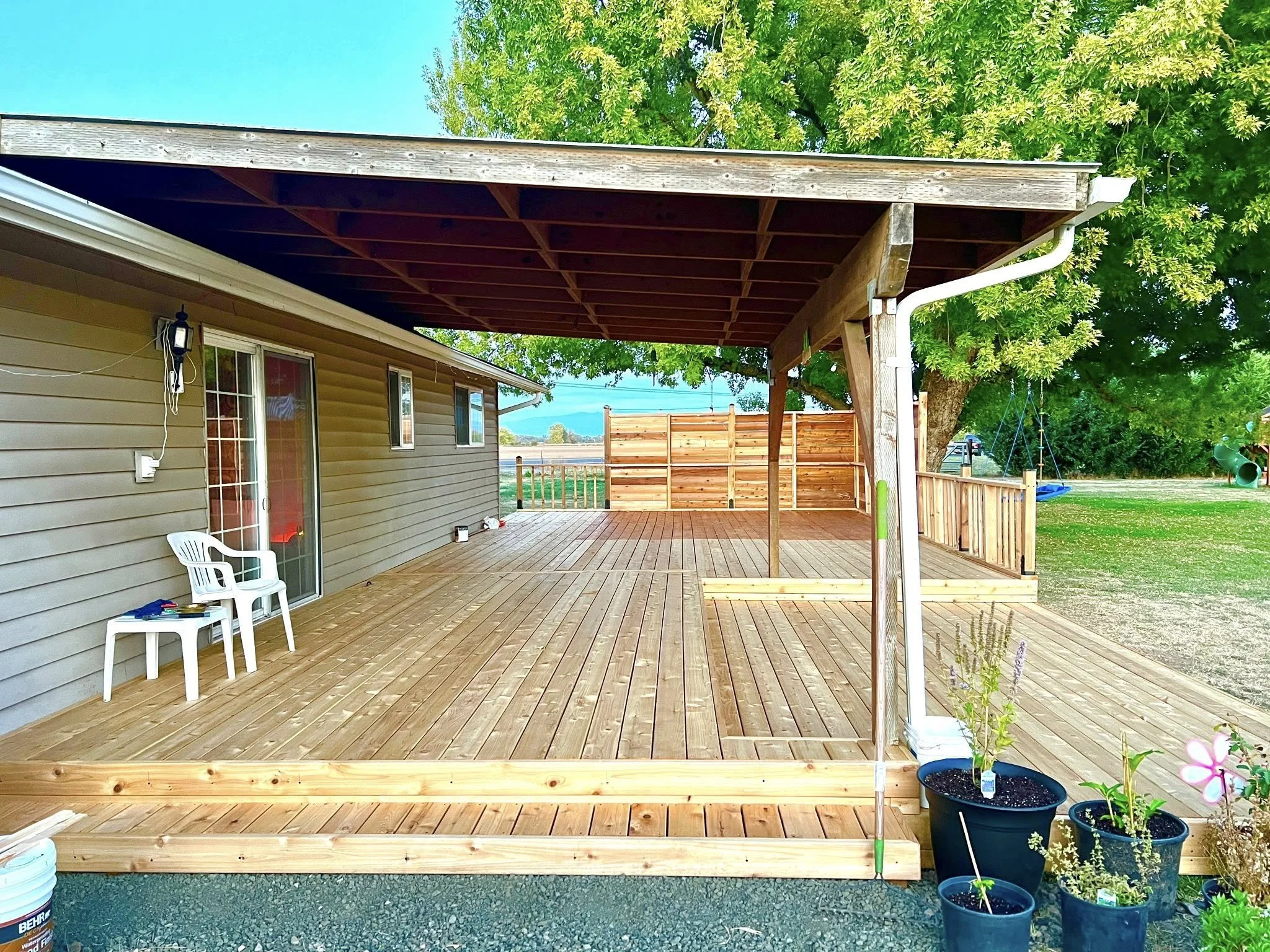 Newly built wooden deck attached to a house, with two white plastic chairs and potted plants, under a covered section with a slanted roof, surrounded by trees and a backyard area.