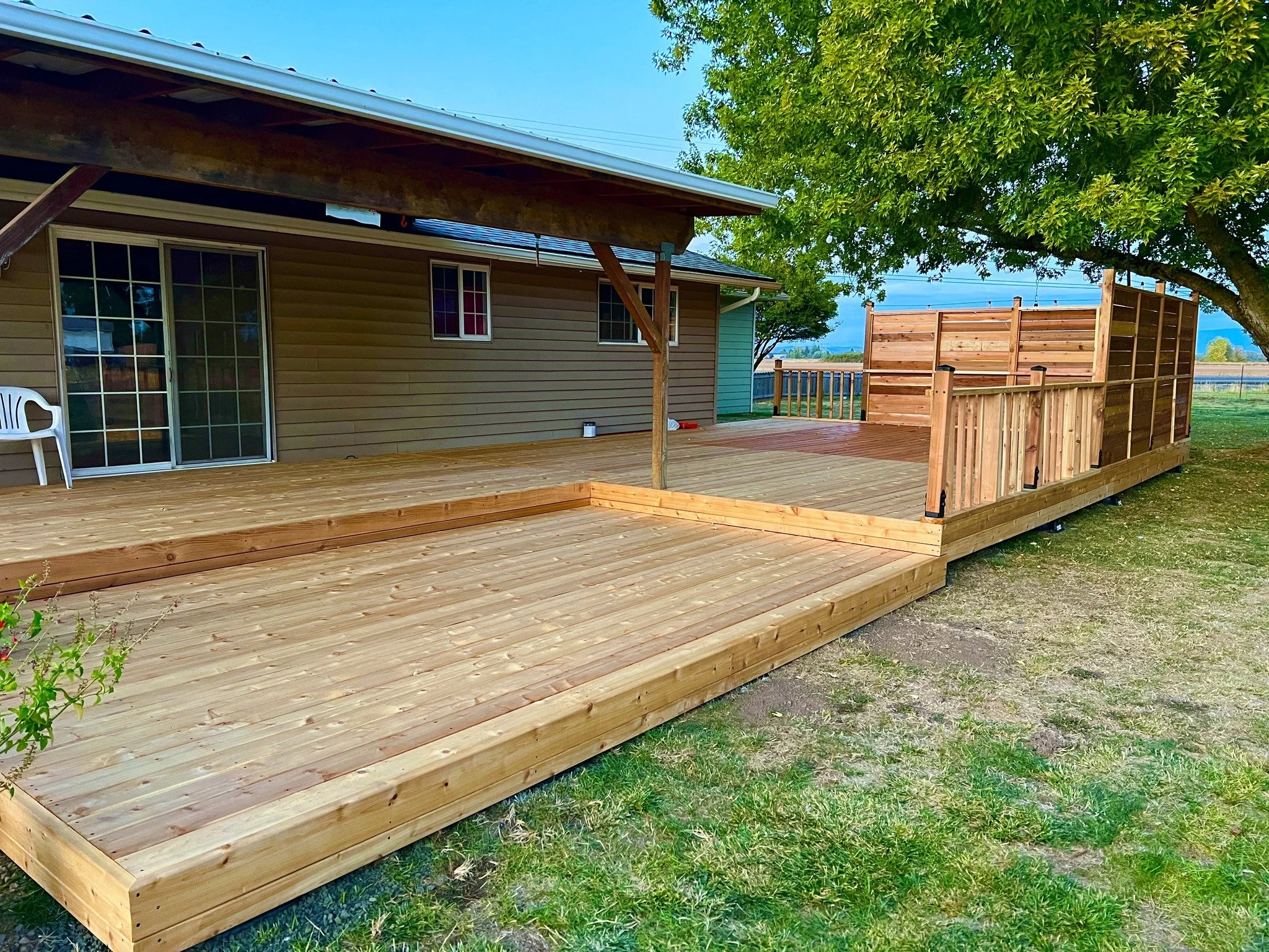 Newly built wooden deck with a sitting area and a wooden privacy fence, attached to a house with beige siding, green shutters, and a large tree on the right side providing shade.