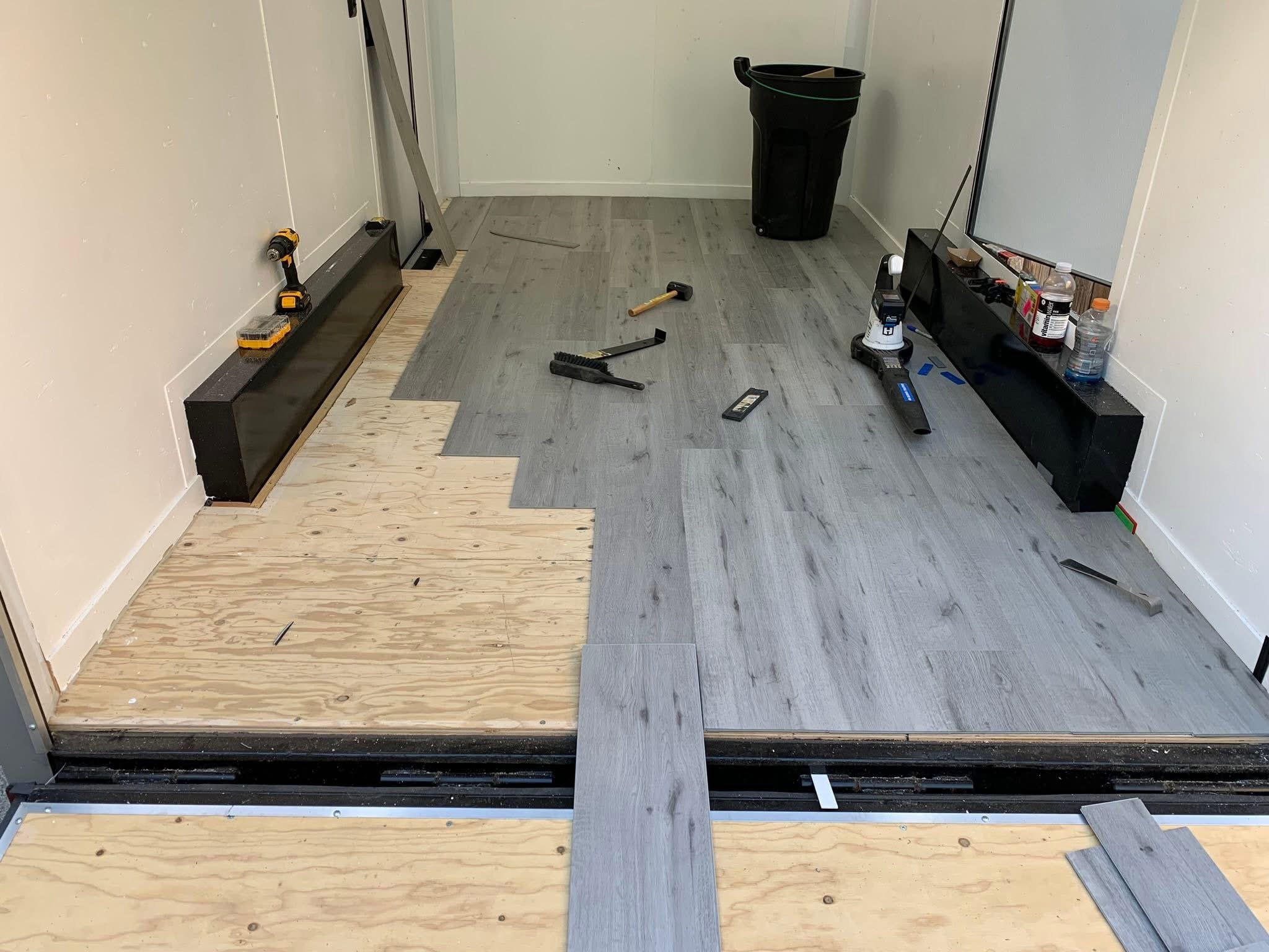 Room undergoing flooring renovation with grey wood laminate flooring partially installed, tools scattered on the floor, a trash bin, and some supplies on a black shelf.