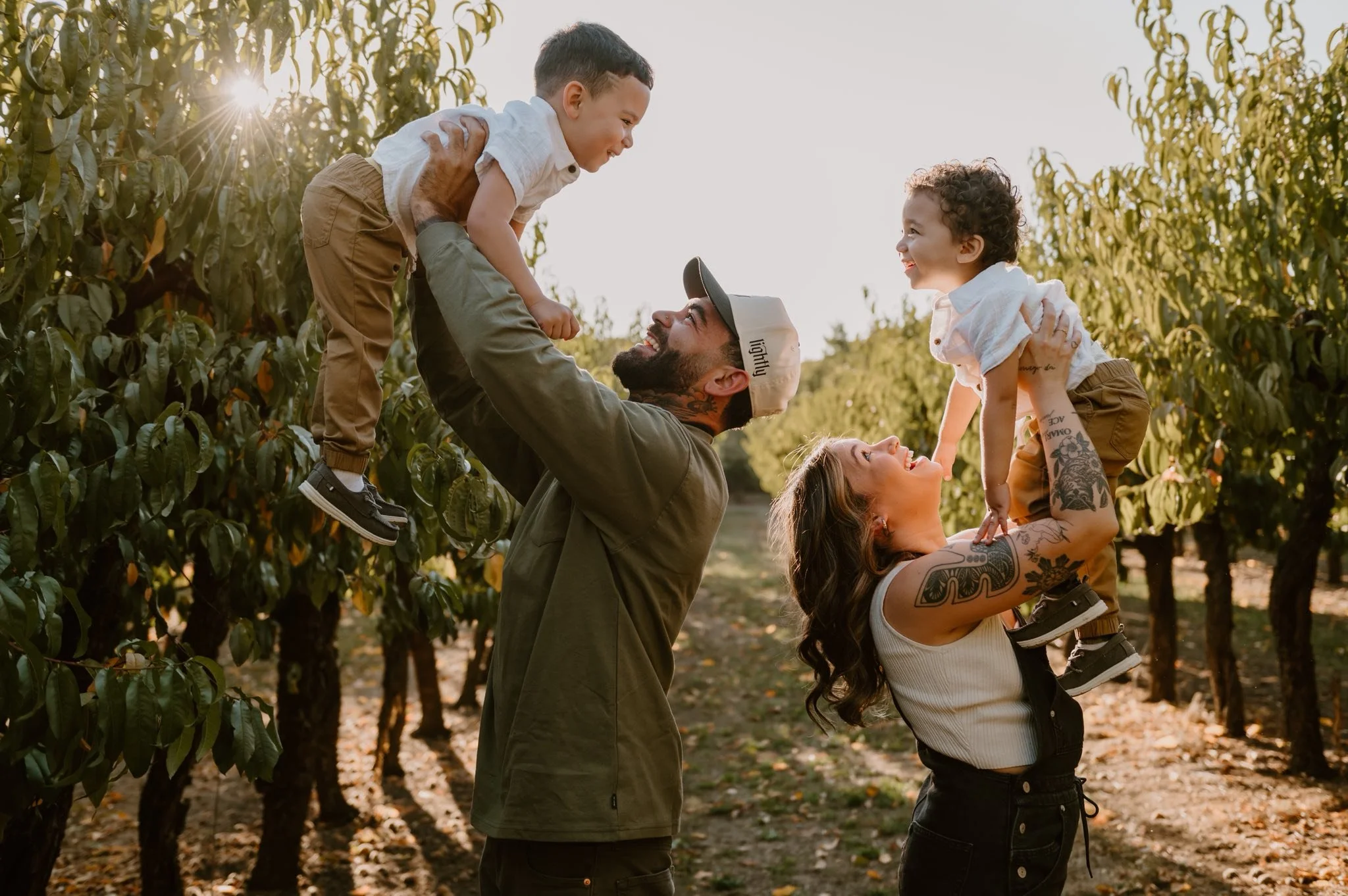 A family of four, consisting of a father, mother, and two young children, enjoying quality time outdoors in an orchard during sunset. The father and mother are lifting their children, a boy and a girl, into the air.