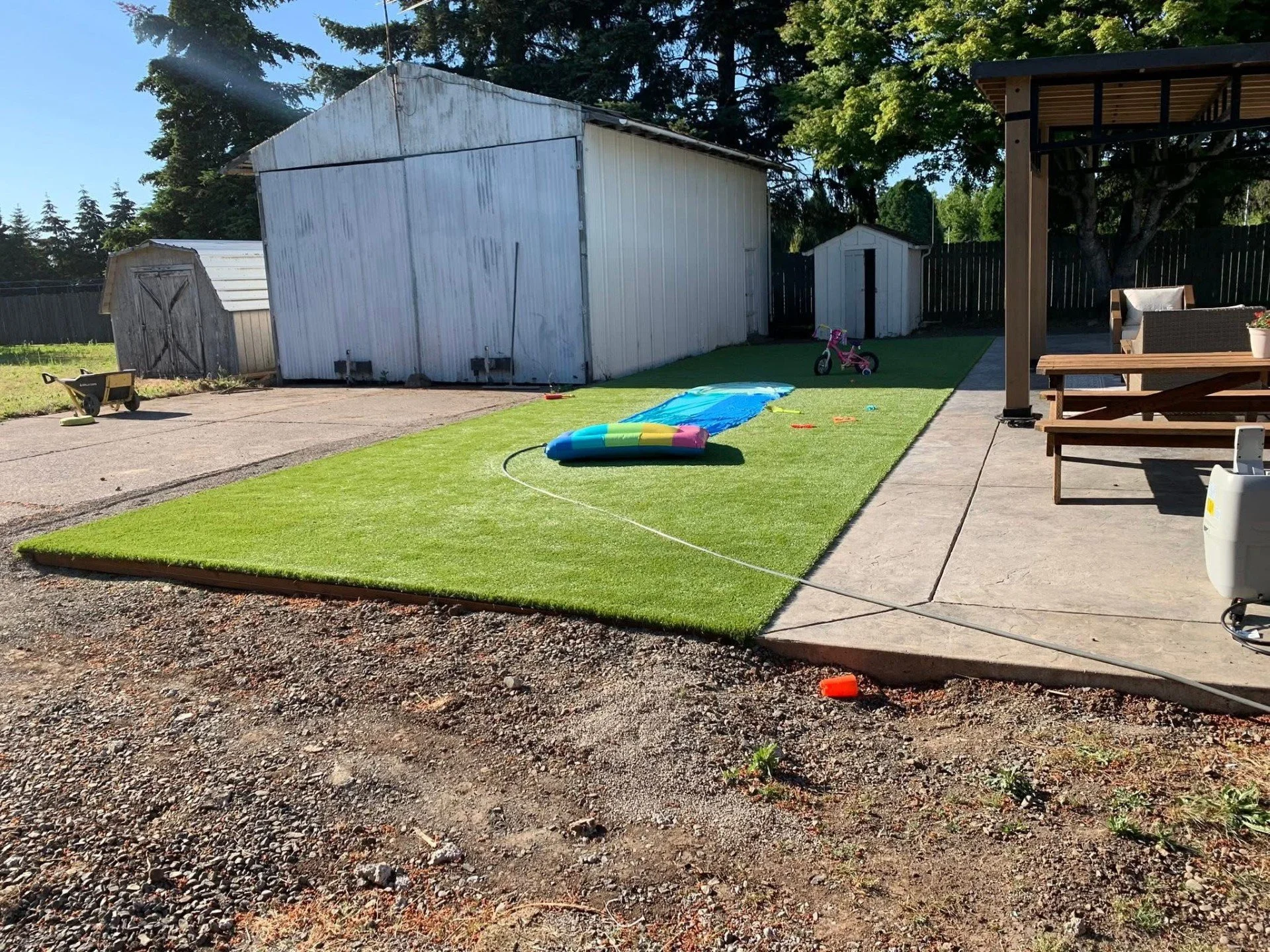 Backyard with a small artificial lawn, children's toys including an inflatable pool, a slide, a toy wheelbarrow, and a pink bicycle, with a concrete patio, outdoor furniture under a gazebo, and shed structures in the background.