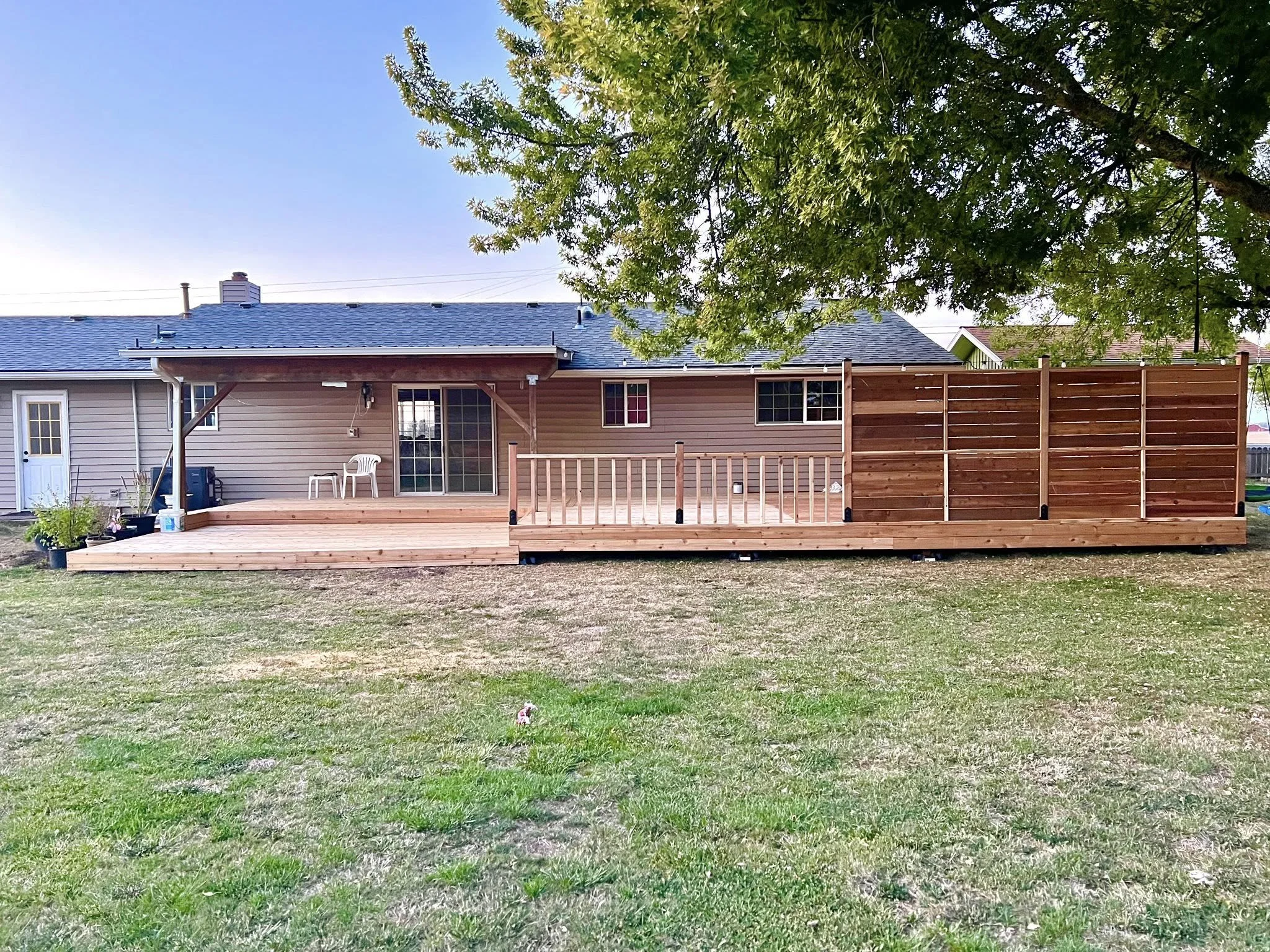 Backyard with a newly built wooden deck attached to a house, featuring a small railing, a white plastic chair, and a sliding glass door. A large tree with green leaves overhangs the deck, and there is a privacy fence on one side. The grass in the yard is patchy.