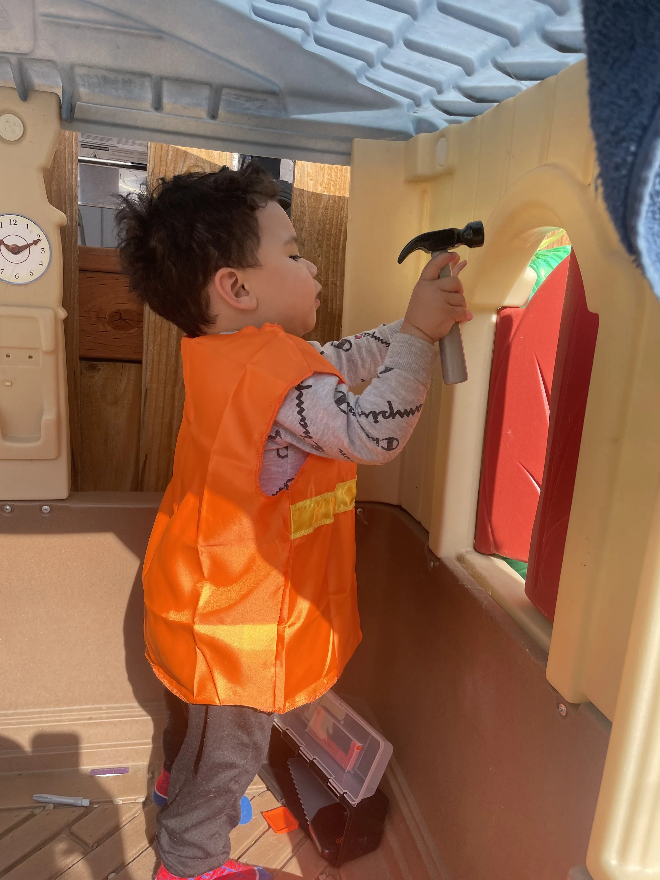 Child wearing an orange safety vest playing with a toy hammer on a play kitchen set.