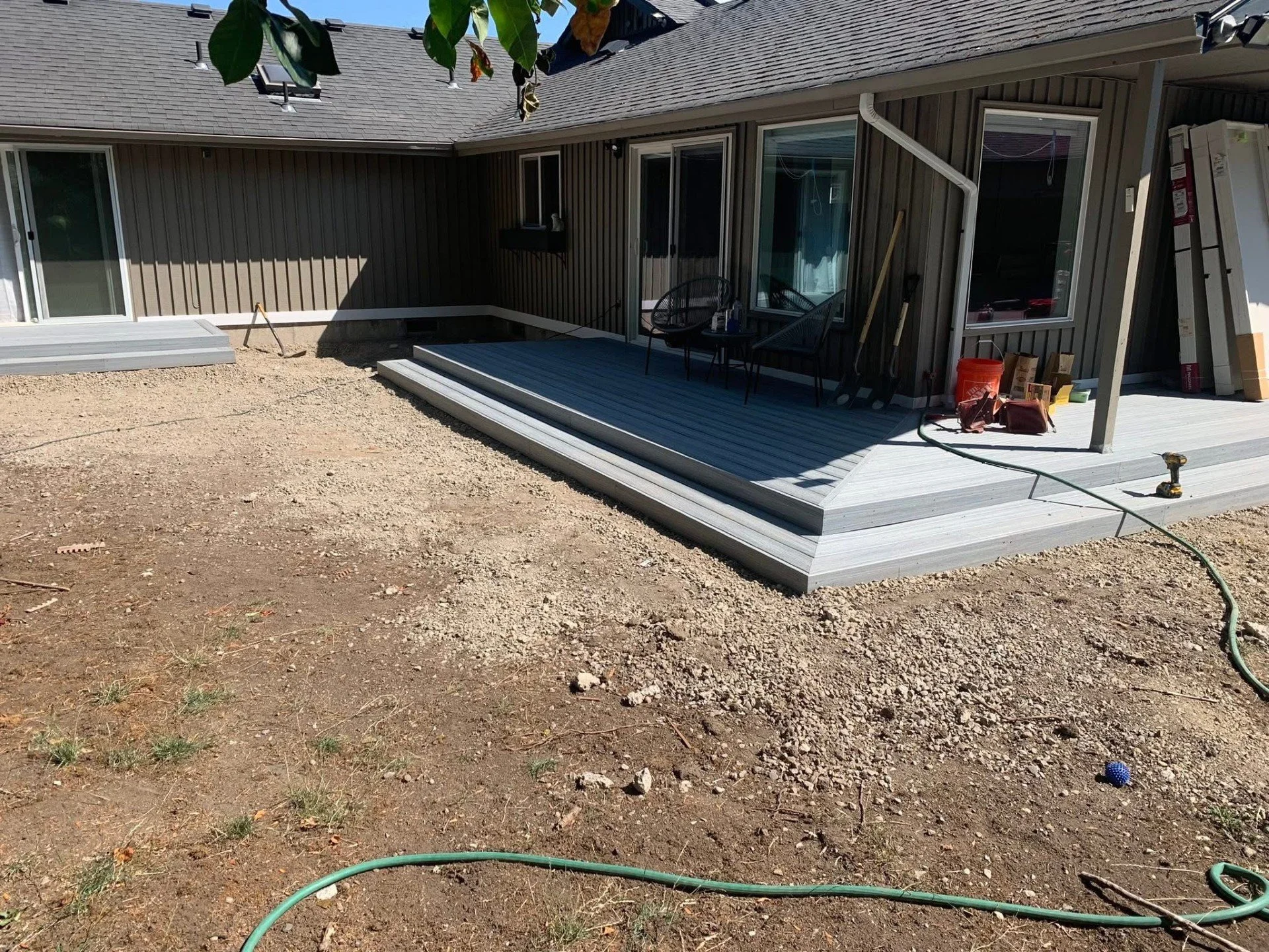 Backyard of a house under construction with a newly built wooden deck, tools, and materials nearby, and bare ground surrounding the deck.
