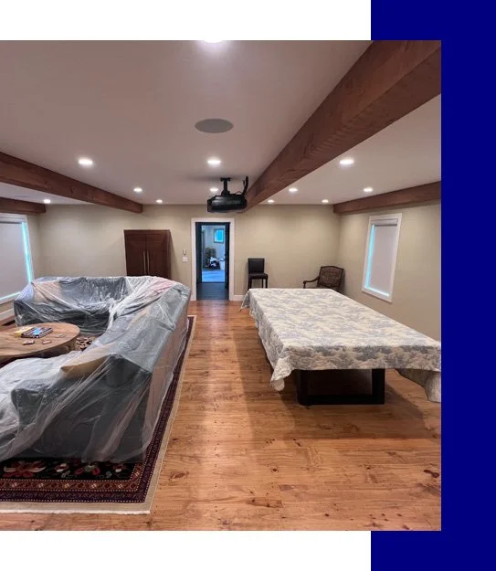 A spacious room with wood flooring, decorated in neutral tones, featuring a covered sofa on a patterned rug, a table, a bed with a floral bedspread, and several chairs. The room has recessed lighting and a ceiling-mounted projector.