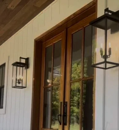 Wooden-framed glass door with black outdoor lanterns on a white wall, greenery reflection visible in the glass.