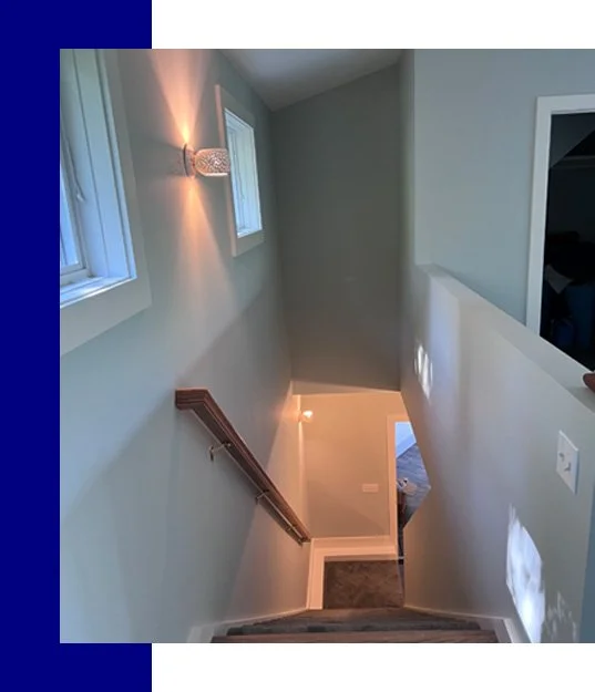 Top view of a staircase from the upper floor looking down, showing light-colored walls, a wooden handrail, two small windows, and a wall-mounted light fixture.