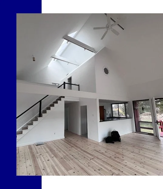 Empty living room with high vaulted ceiling, skylight, ceiling fan, staircase, and large windows.