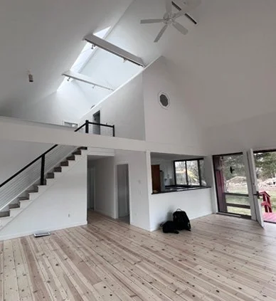 Interior view of a spacious, modern, open-plan living area with high ceilings, large windows, and hardwood flooring, featuring a staircase with black railing and a ceiling fan.