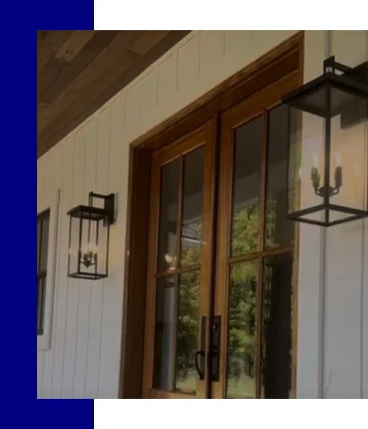 Exterior view of a house showing a wooden door with glass panels and two black metal outdoor lanterns on the white siding wall.