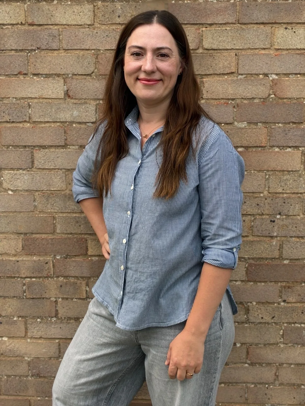 A white woman in her 40s with a blue shirt, grey jeans and long brown hair, smiling.