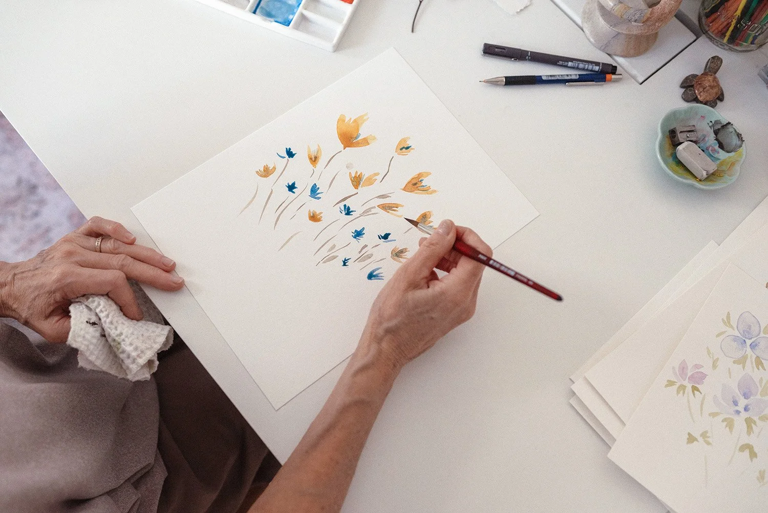 top down view of woman painting orange and blue water colour flowers on desktop