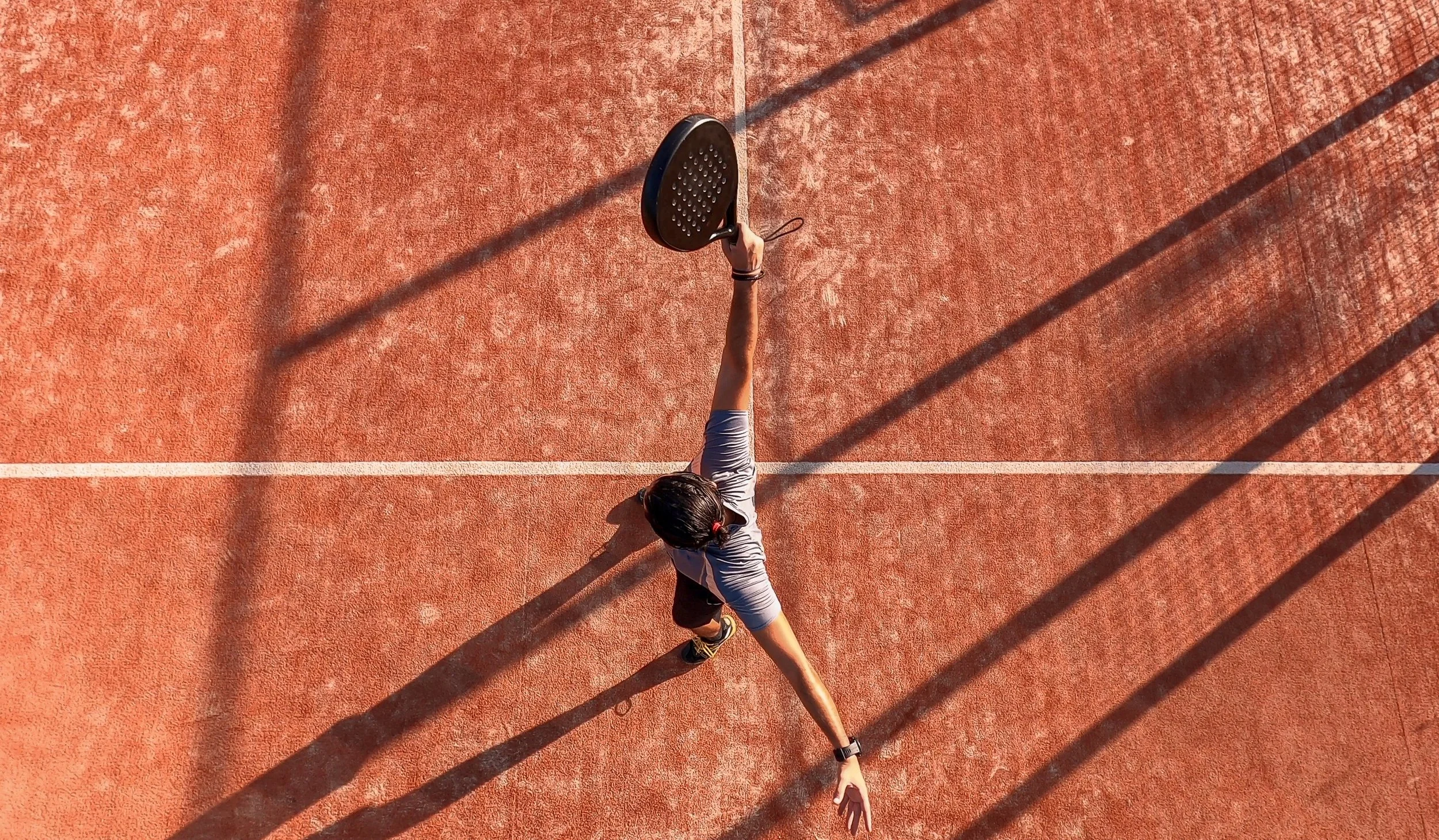Person playing tennis on an outdoor court, reaching for the ball with a paddle, with long shadows cast on the red surface.