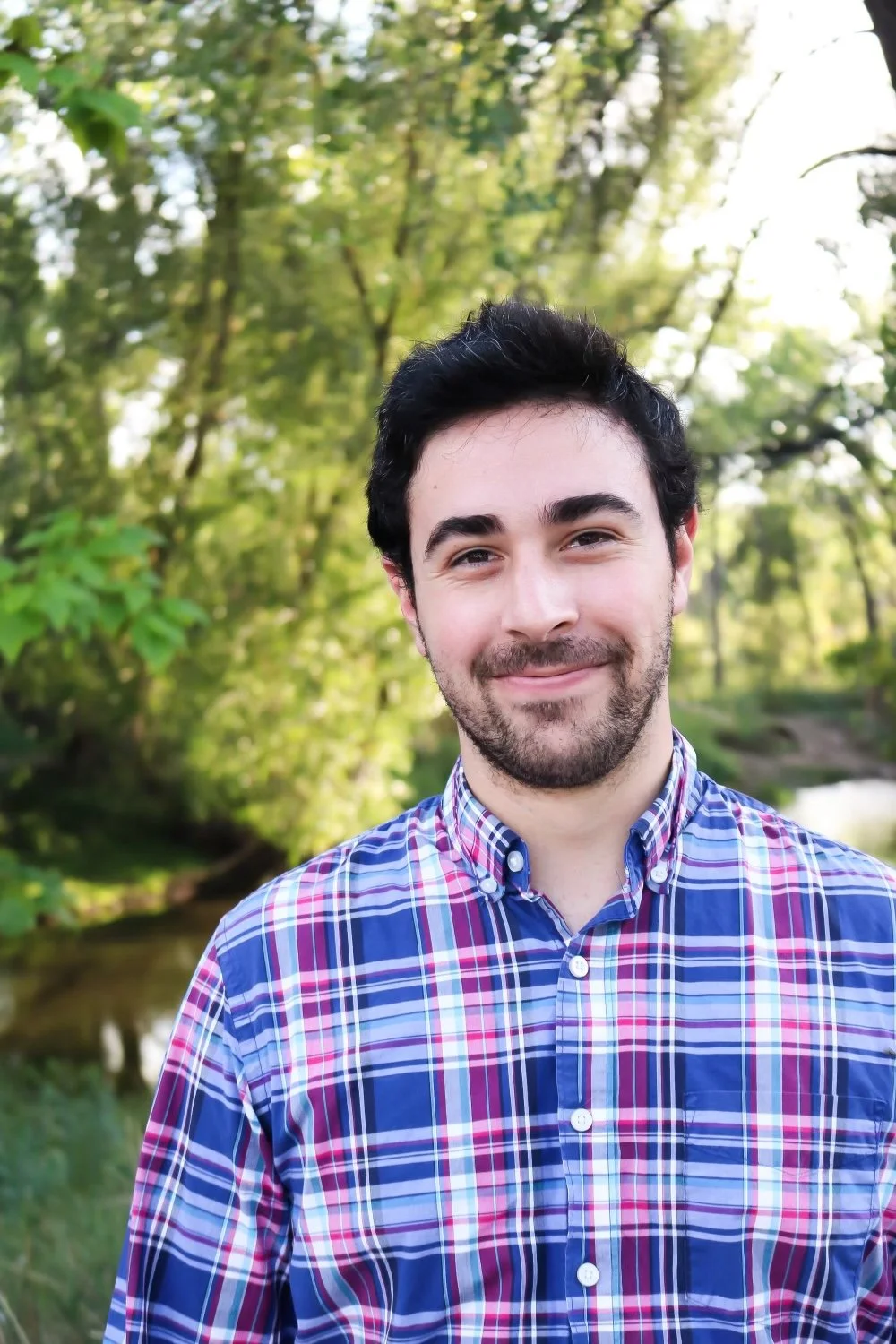 A young man with dark hair, a beard, and mustache smiling outdoors in front of a wooded area with green trees and a small body of water.