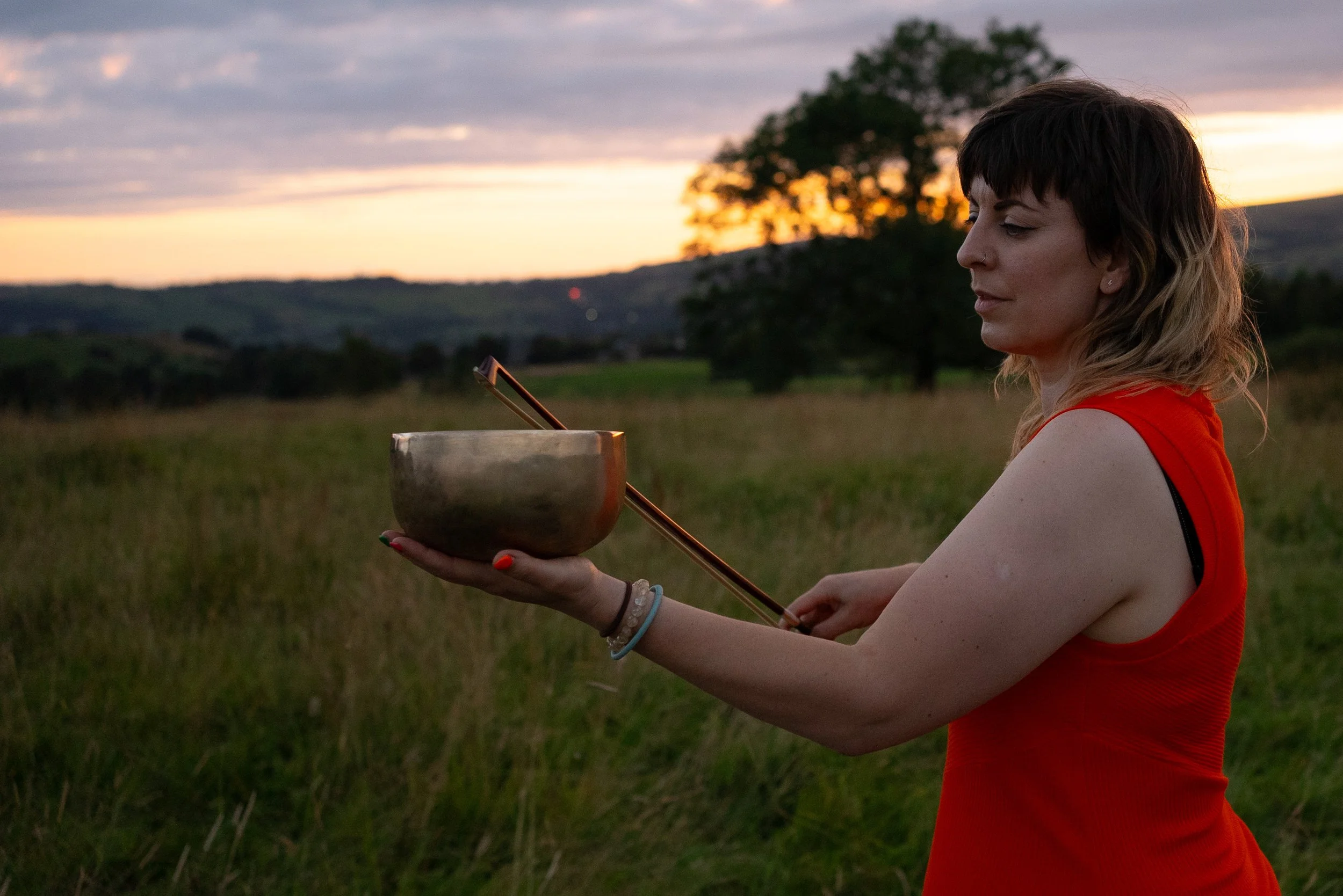 A woman in a red tank top holding a singing bowl and a mallet outdoors during sunset.