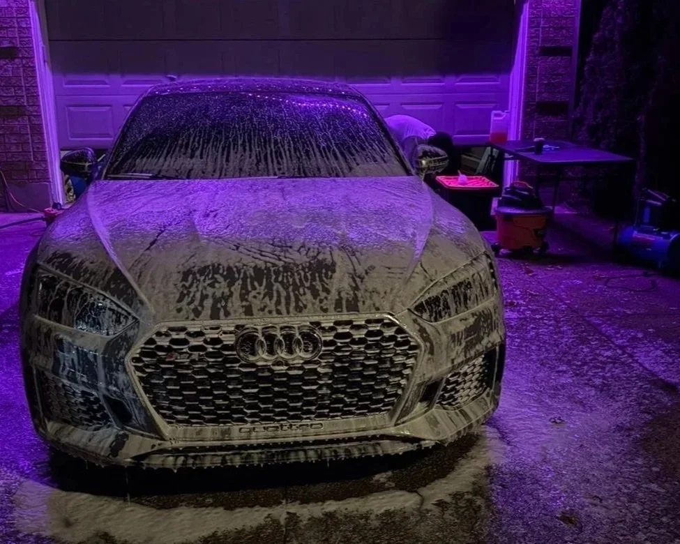 An Audi car covered in soap suds during a car wash at night in a garage illuminated with purple lighting.