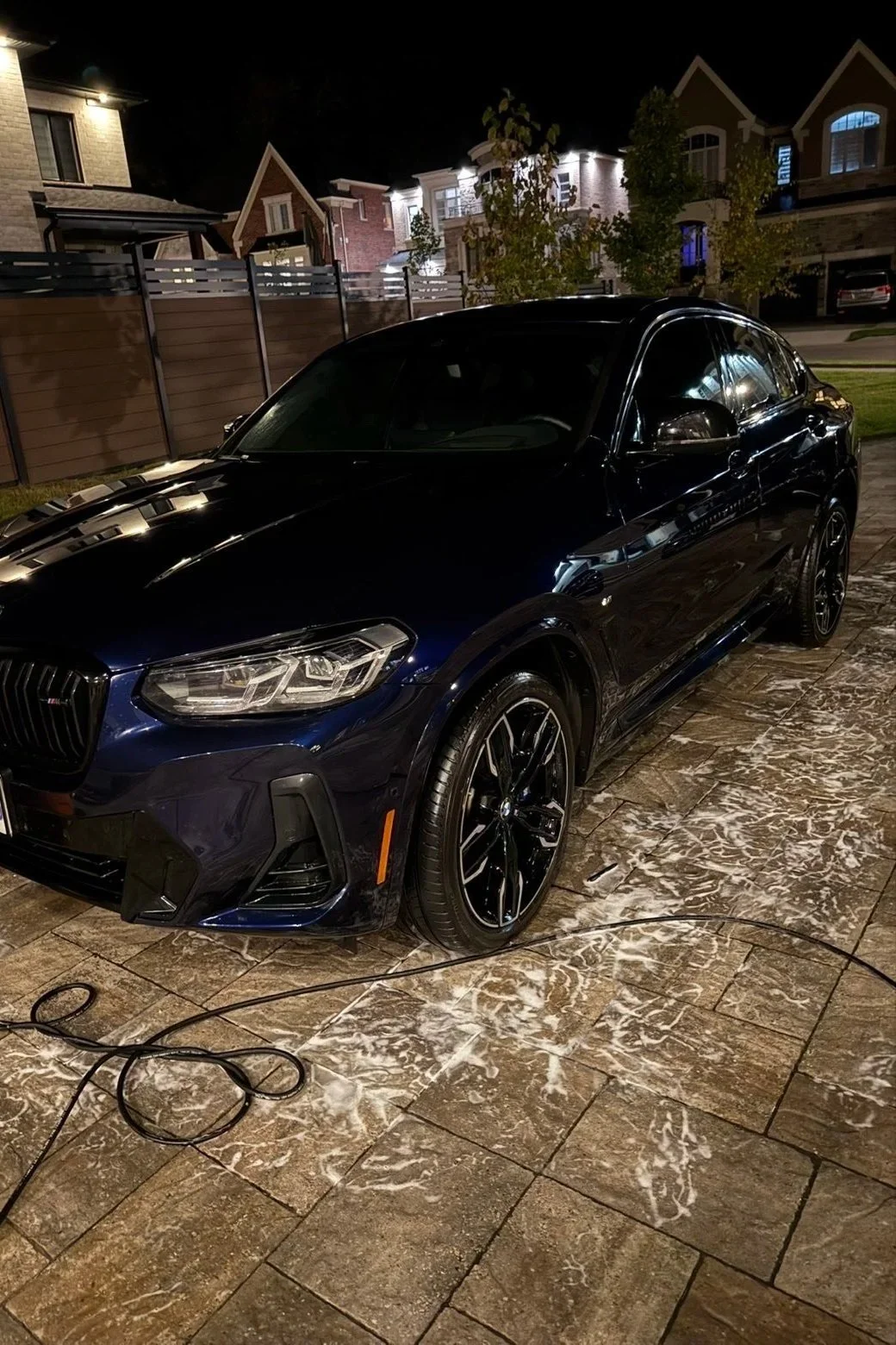 A black sports sedan parked on a wet stone patio at night, with a suburban neighborhood in the background.