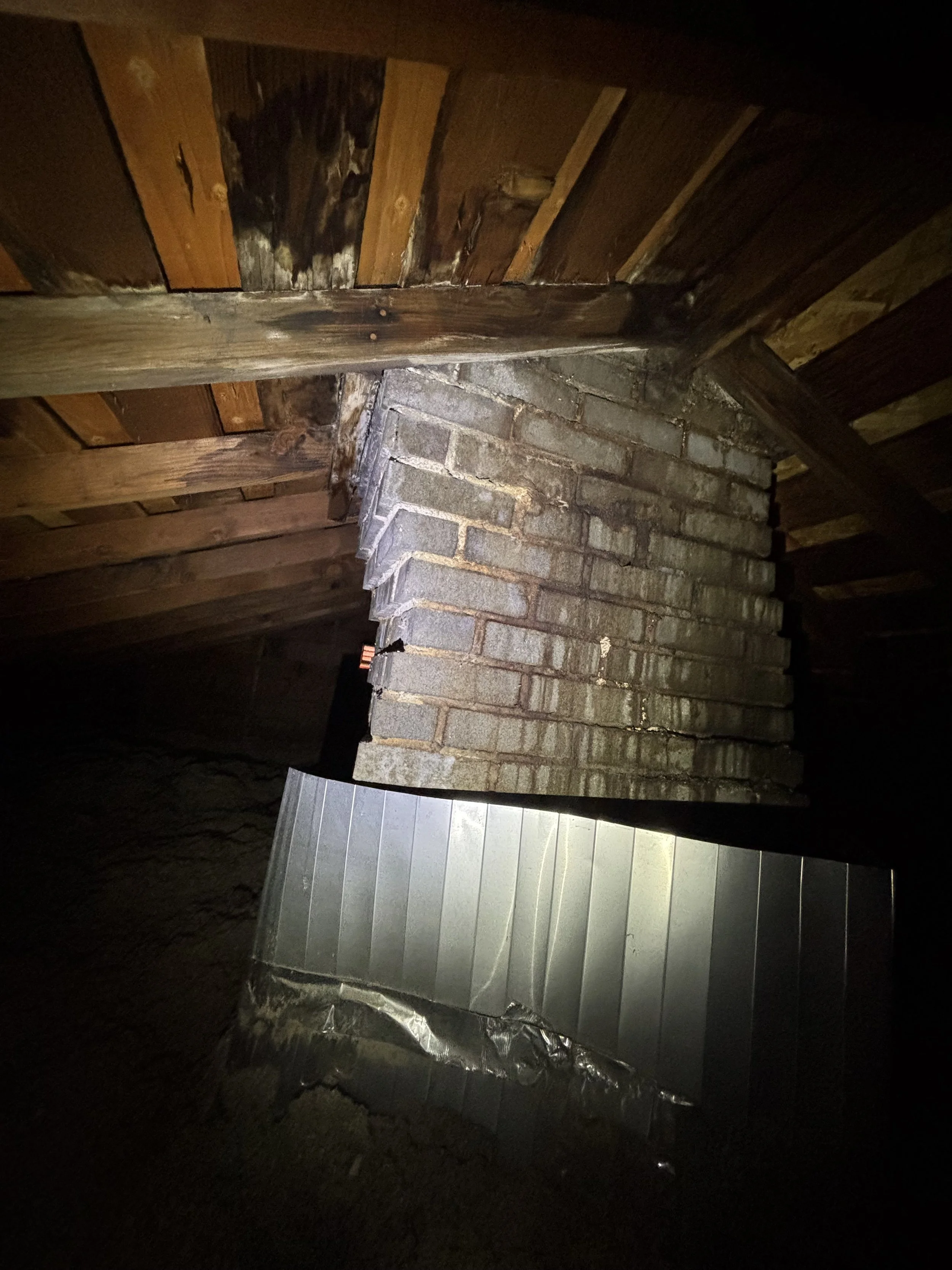 View of a brick chimney in an attic with a wooden roof and a metal sheet leaning against the wall.