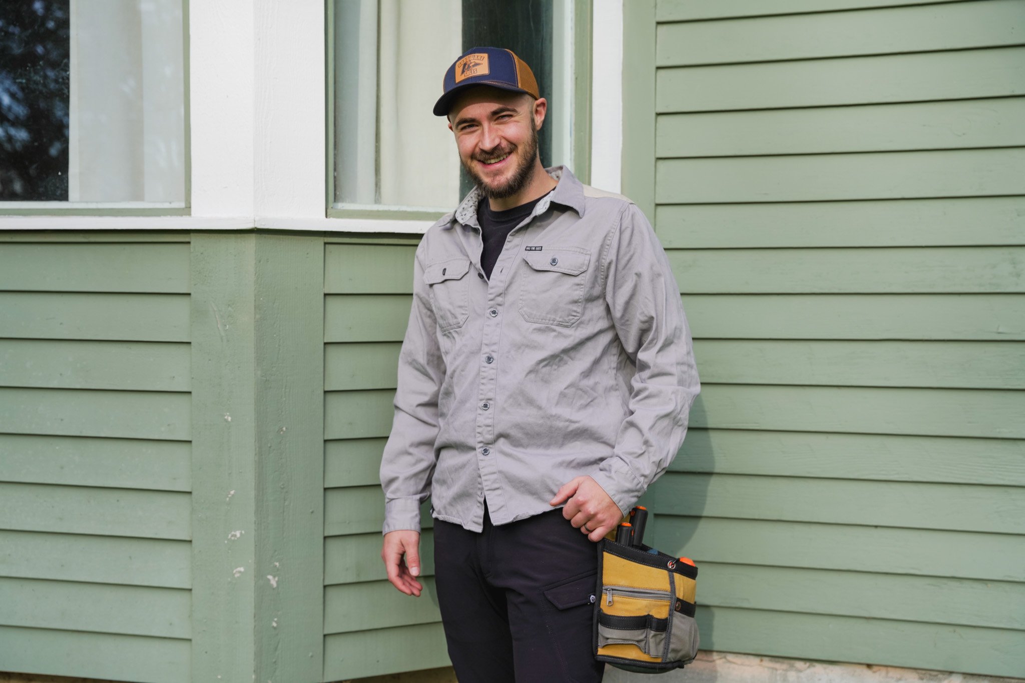 A smiling man standing by a green house, wearing a gray work shirt, black pants, a black and yellow tool belt, and a baseball cap.