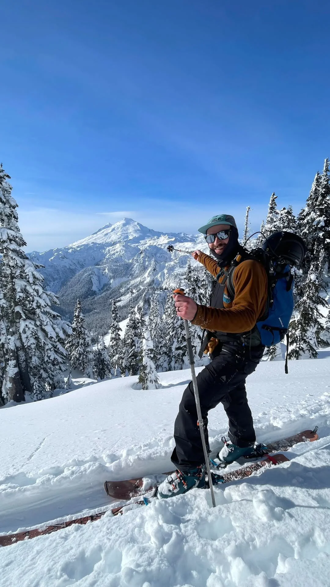A man in winter gear with sunglasses, pointing at a snowy mountain landscape with trees covered in snow.