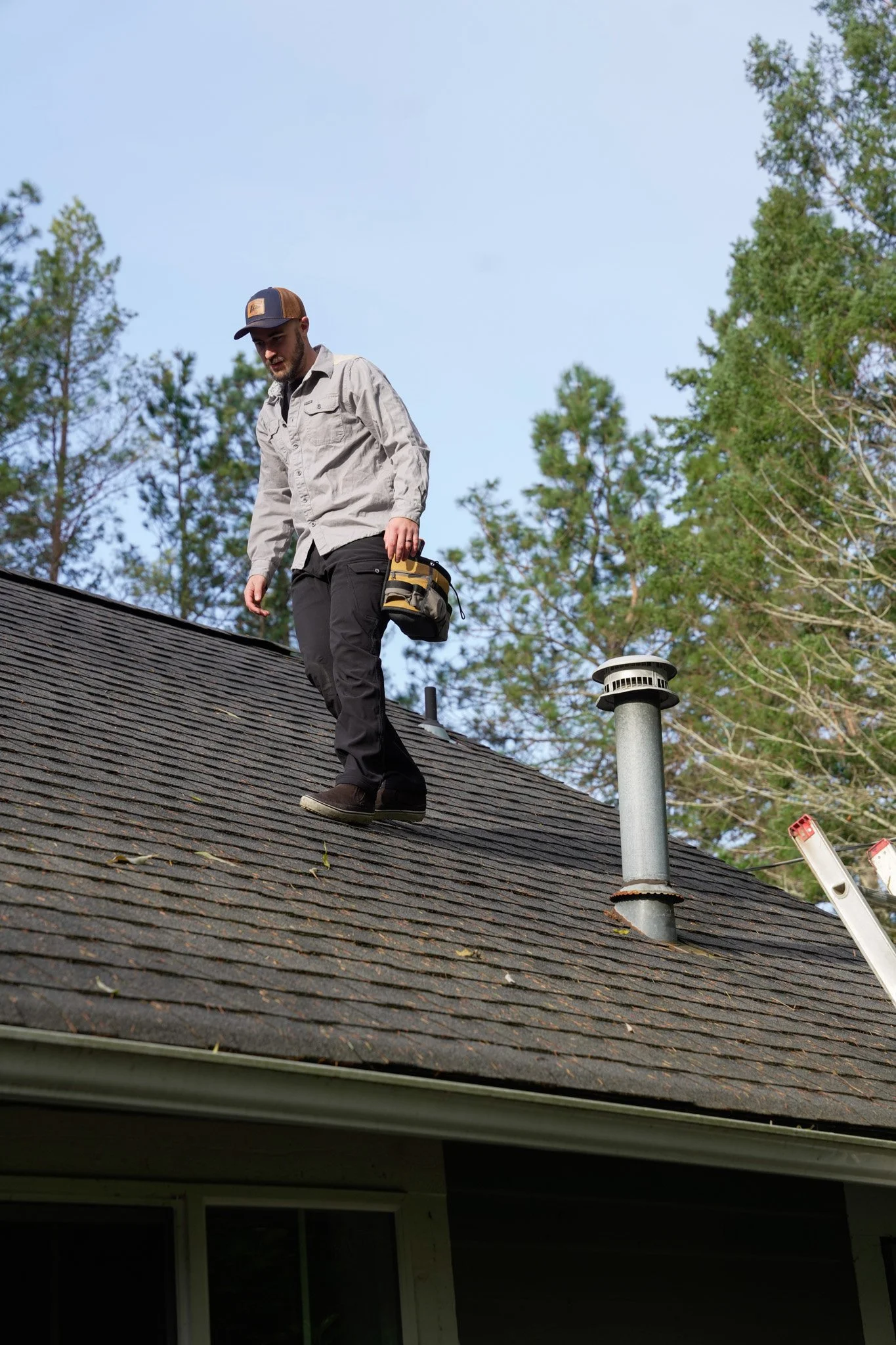 A man walking on a roof with shingles, wearing a cap, a light gray jacket, black pants, and holding a tool bag, surrounded by trees and a ladder.