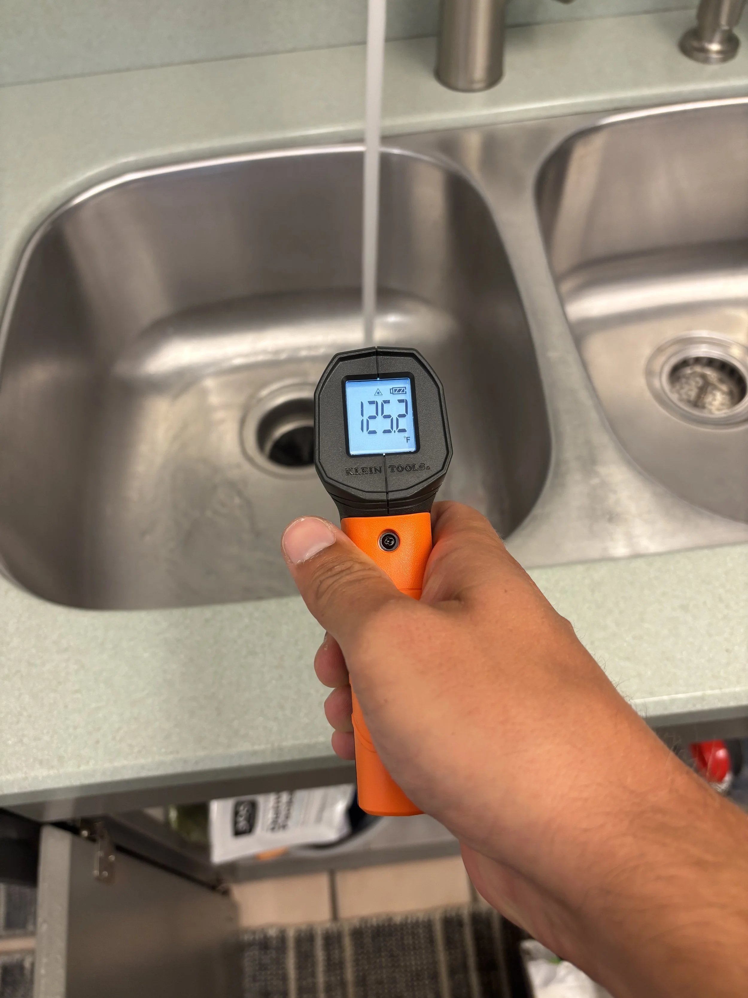 A hand holding a digital infrared thermometer above a kitchen sink, showing a water temperature of 125.2 degrees Fahrenheit.