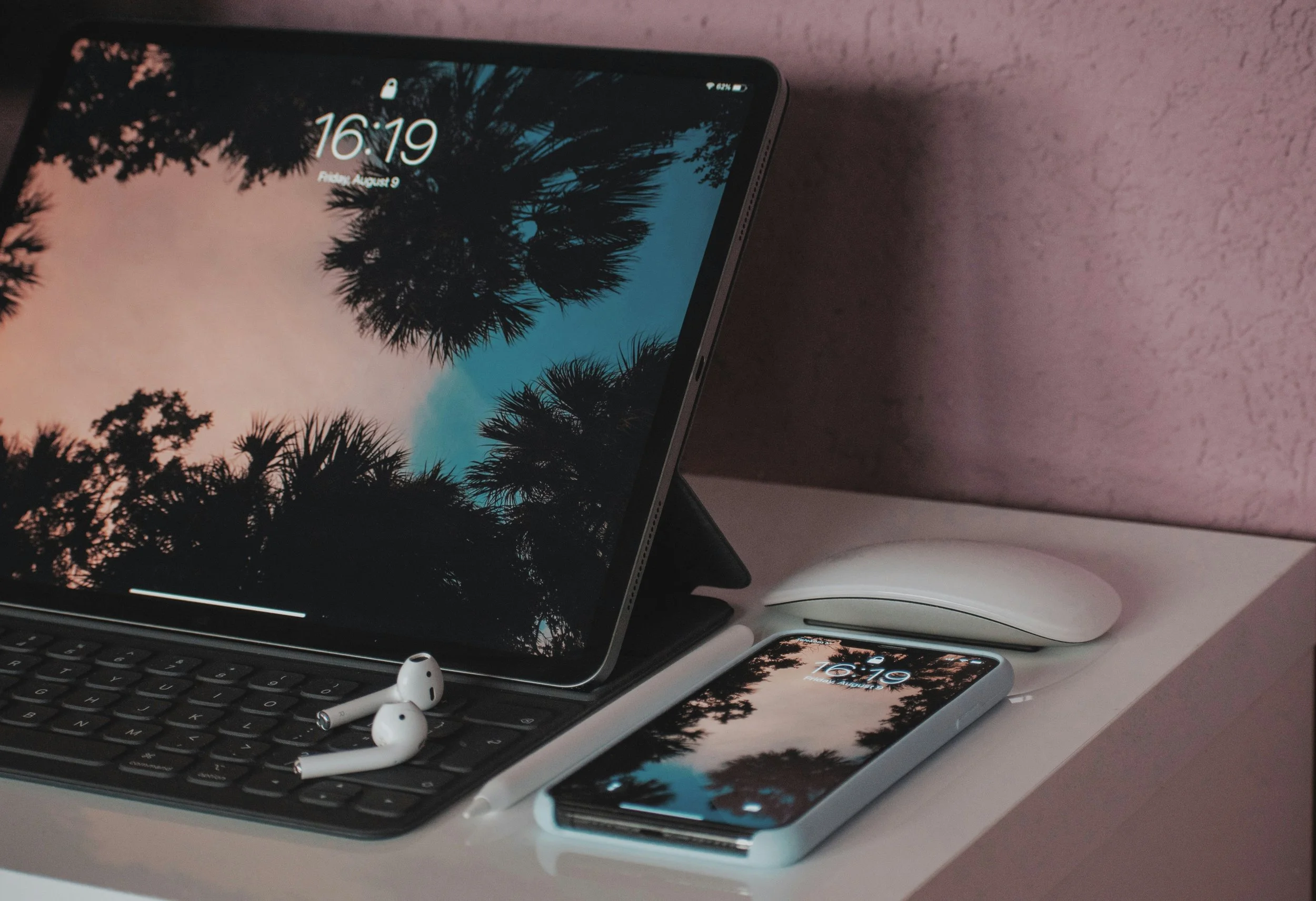 A tablet on a keyboard stand displaying a nature scene with silhouetted trees against a colorful sky, a pair of wireless earbuds on the keyboard, a smartphone with a similar nature scene on the screen, and a wireless mouse on a white desk against a pink wall.