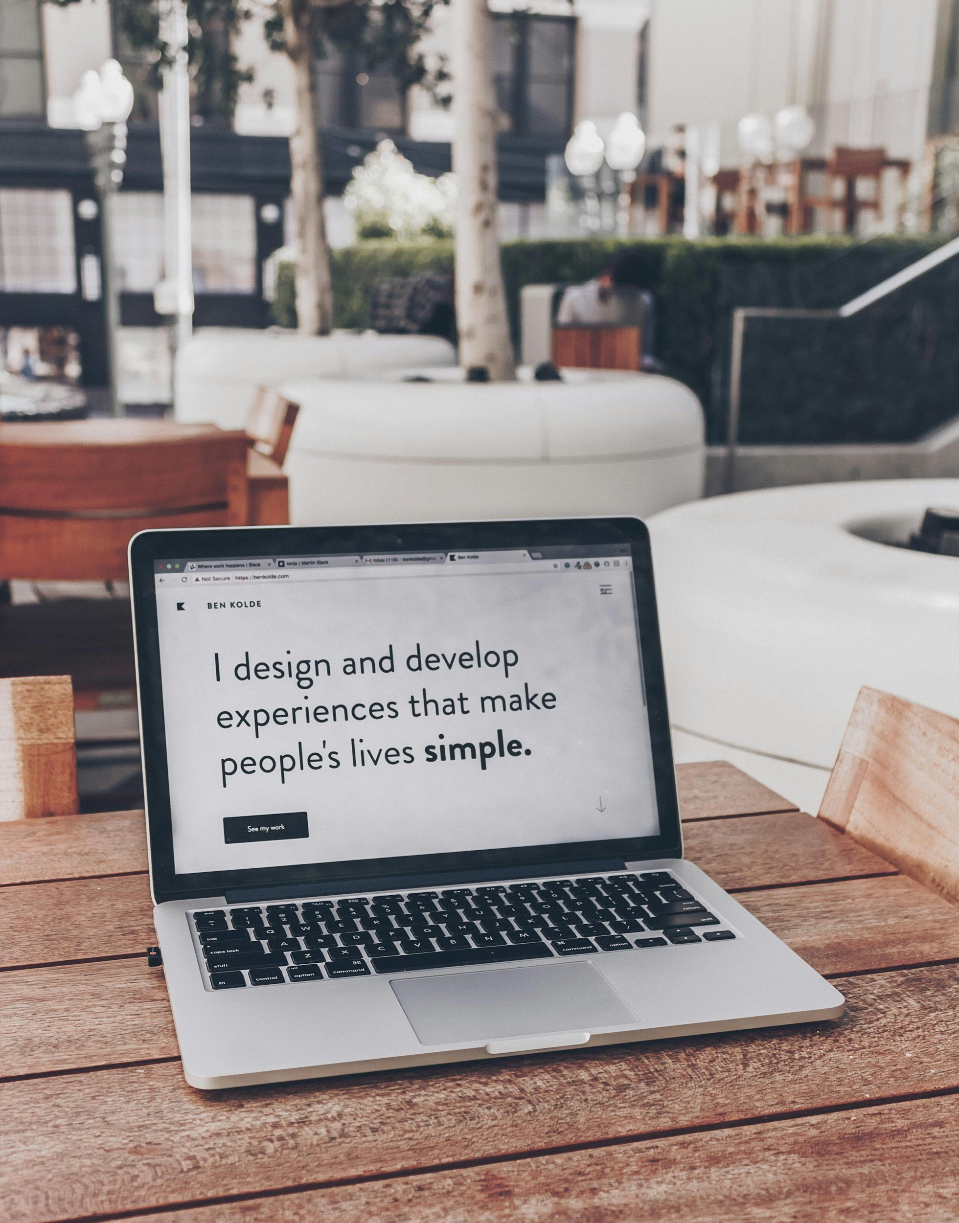 Open laptop on wooden table displaying a website with the text 'I design and develop experiences that make people's lives simple.' in a modern office or lounge setting.