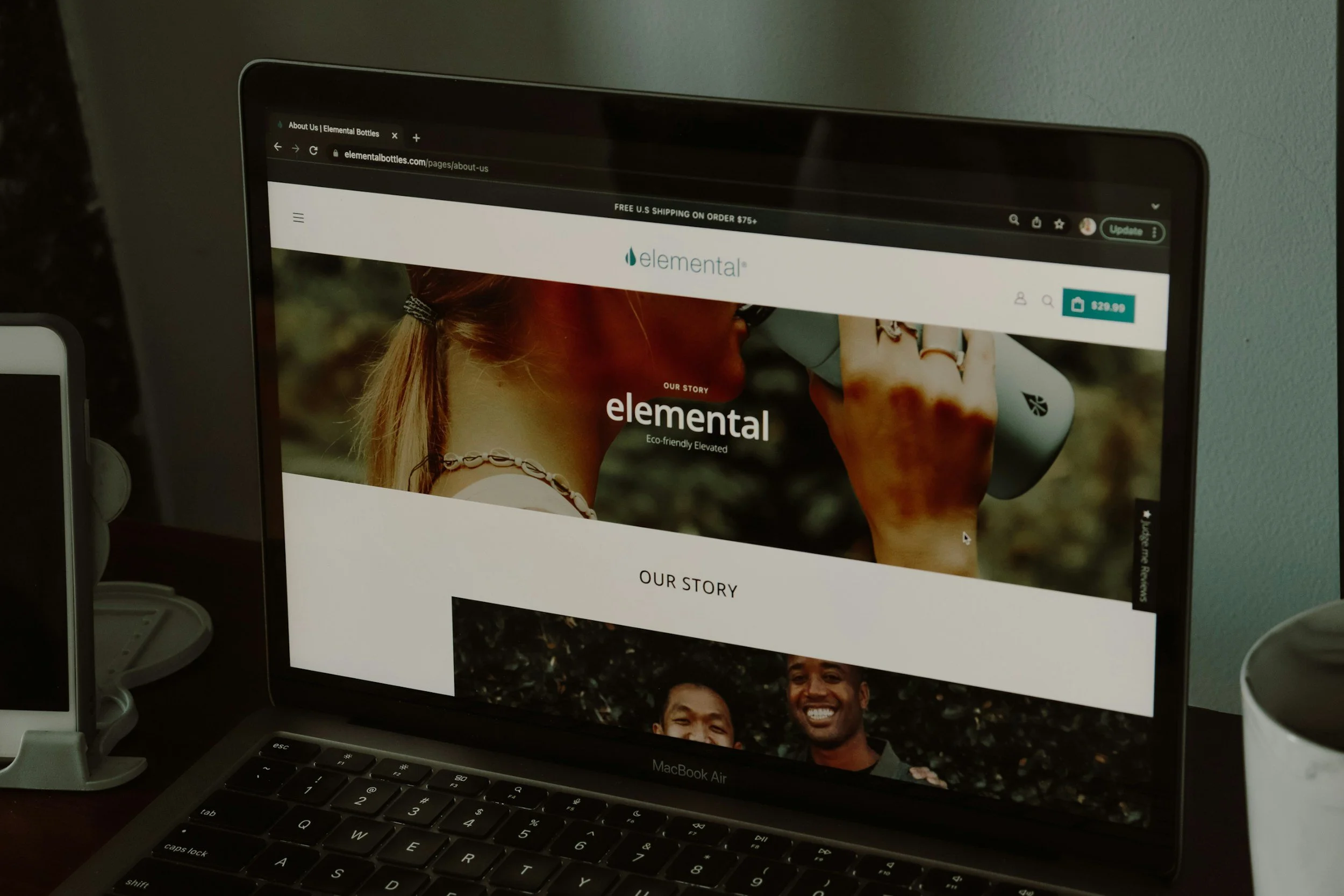 A laptop displaying the webpage for Elemental, an eco-friendly bottled water company, on a desk with a white device to the left and a white cup to the right.
