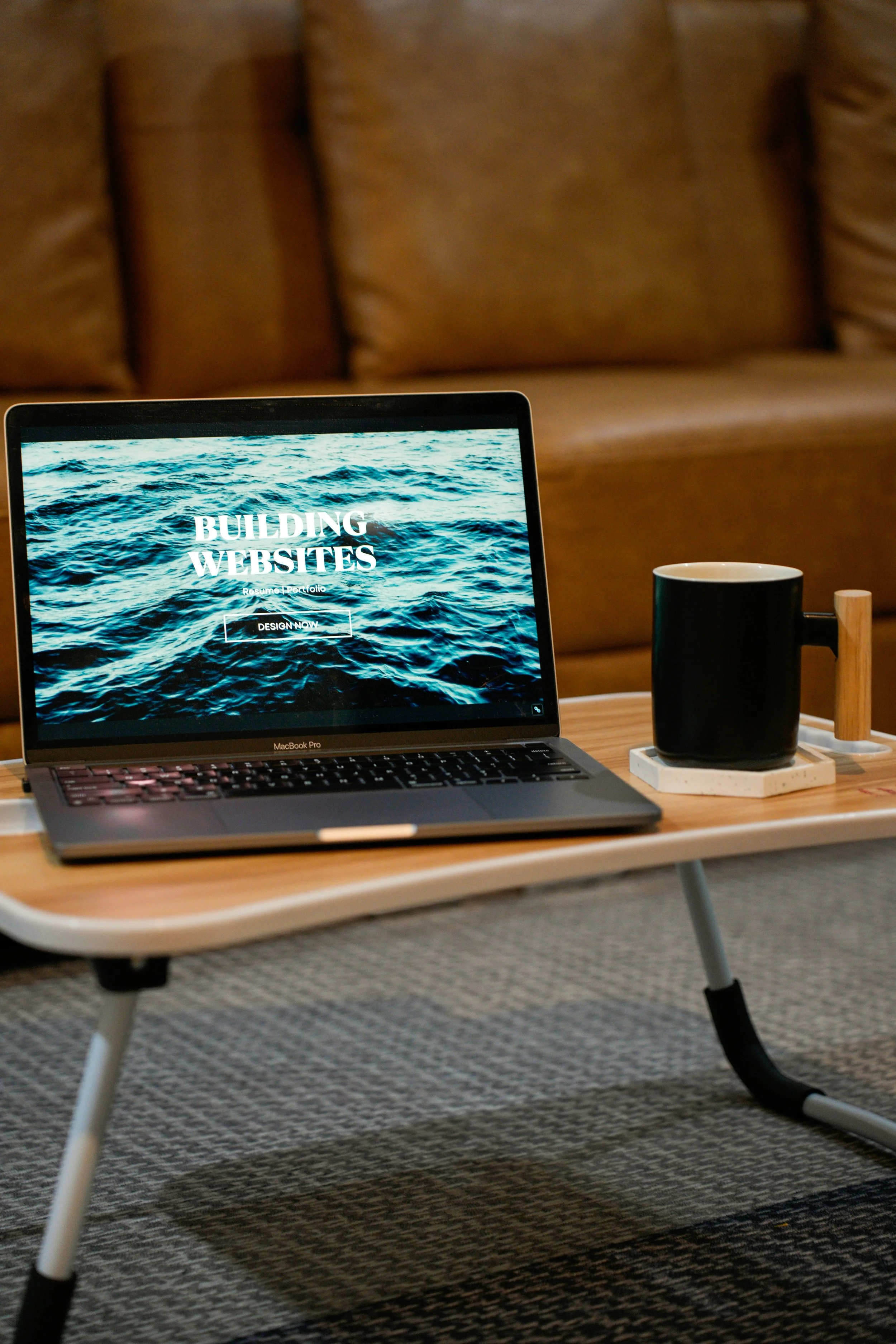 A laptop on a wooden table displaying a website titled 'Building Websites' with a background of ocean waves, next to a black mug on a coaster and a small wooden object, in front of a brown sofa.