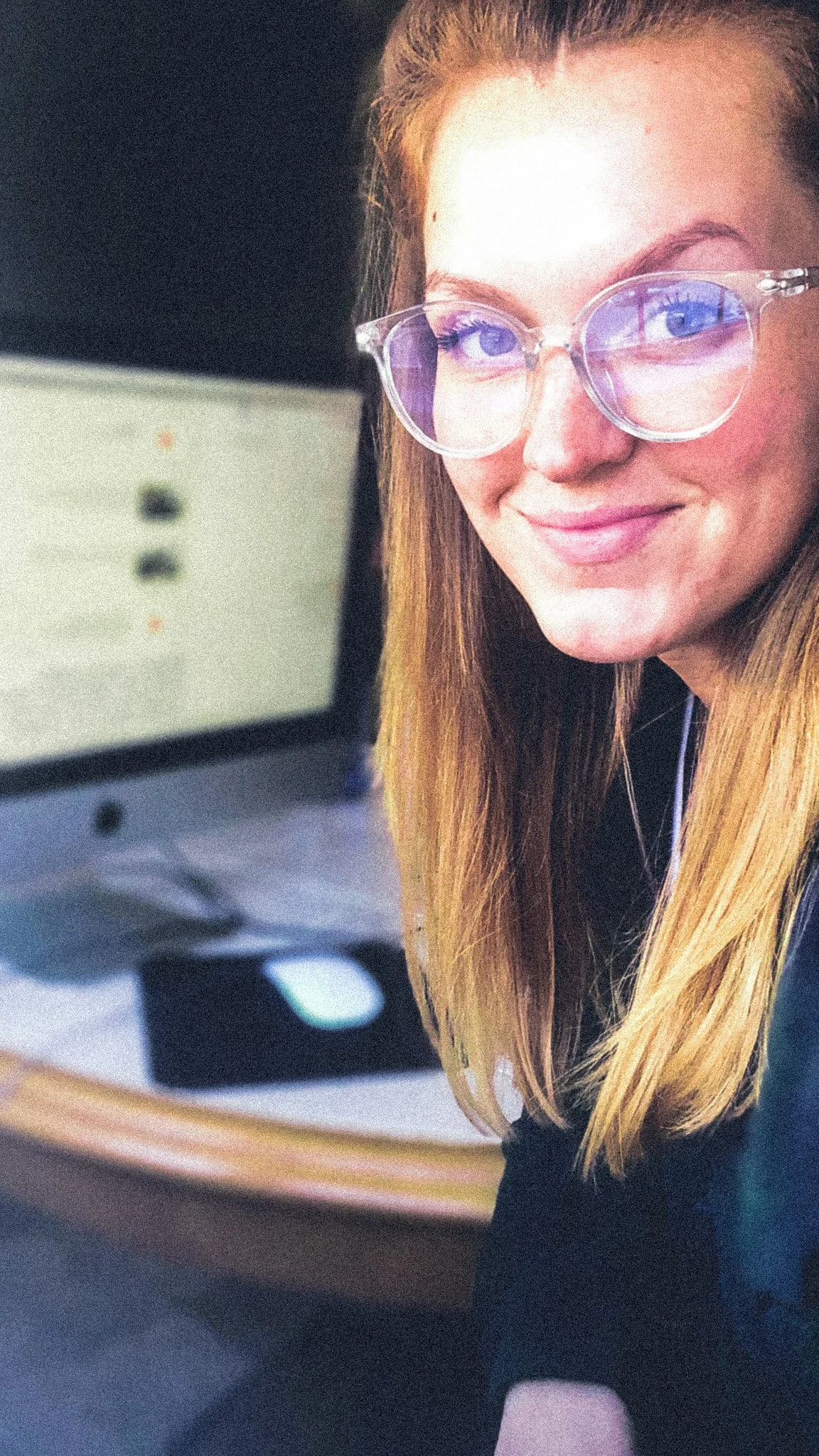 A young woman with blonde hair, wearing clear glasses with purple-tinted lenses, smiling at the camera in front of a computer monitor.