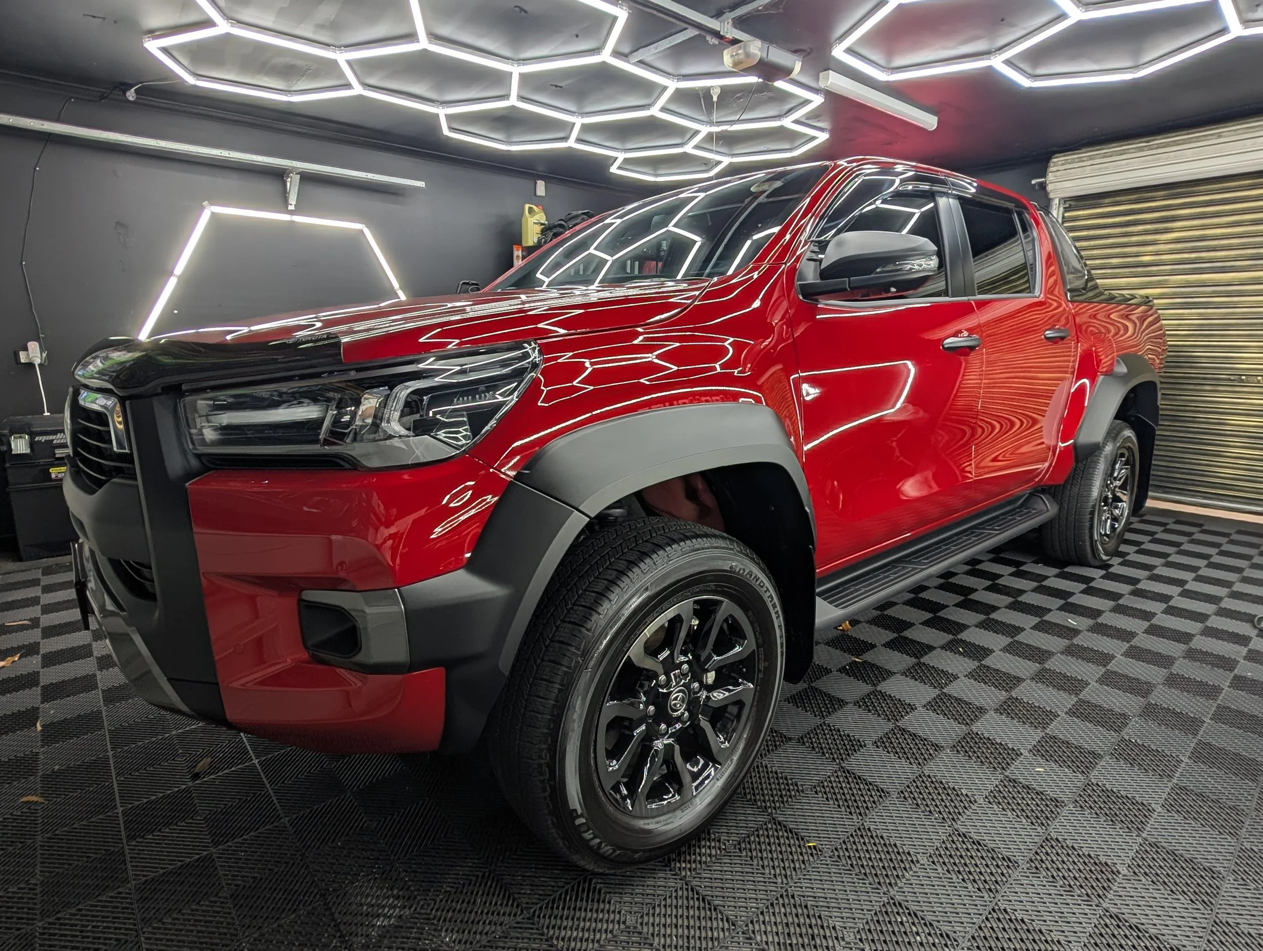 Red pickup truck parked in a garage with black walls and hexagon-shaped ceiling lights.