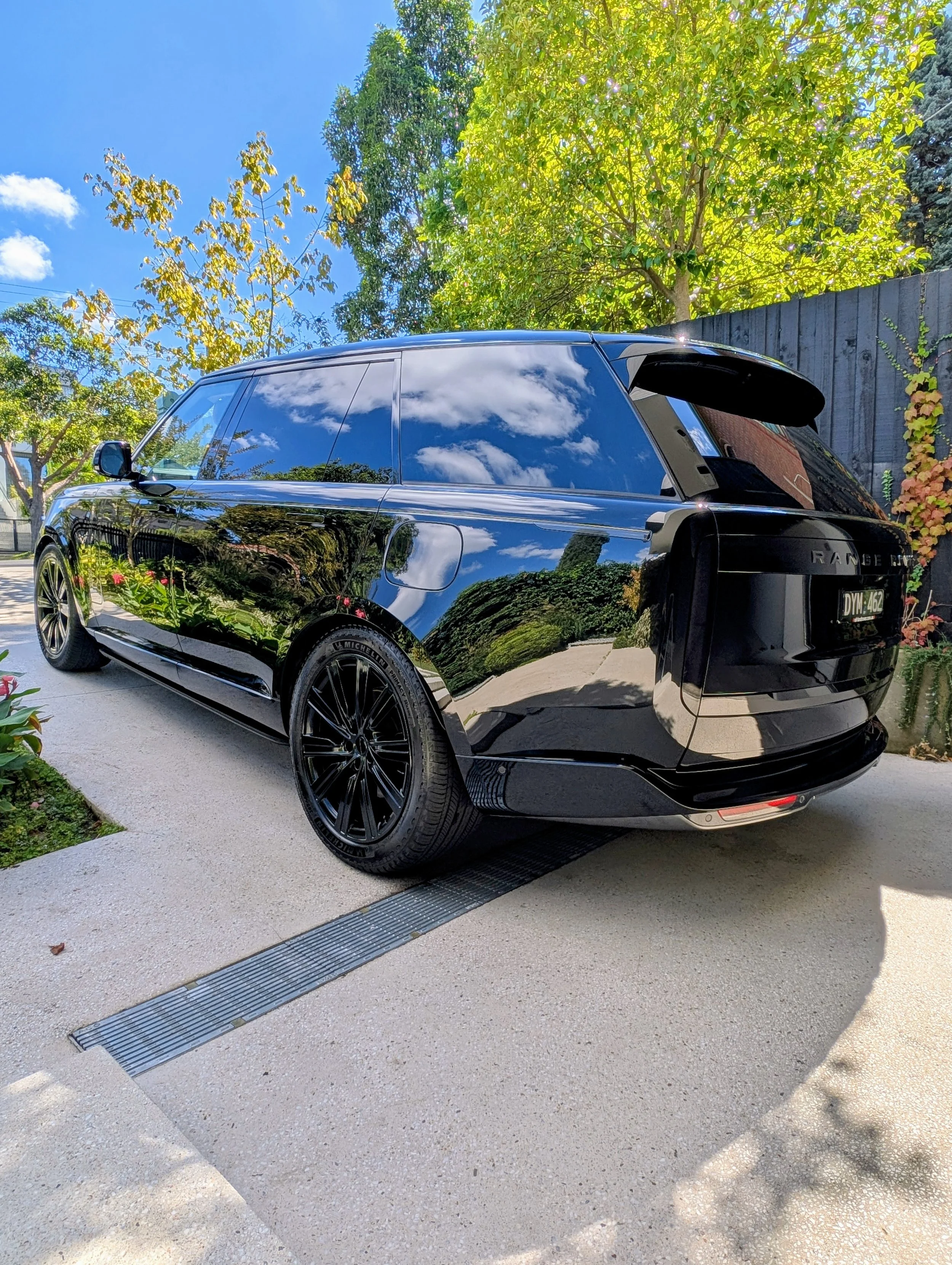 Black Range Rover SUV parked on a driveway, reflecting trees, sky, and clouds on its shiny surface, with green trees and a wooden fence in the background.