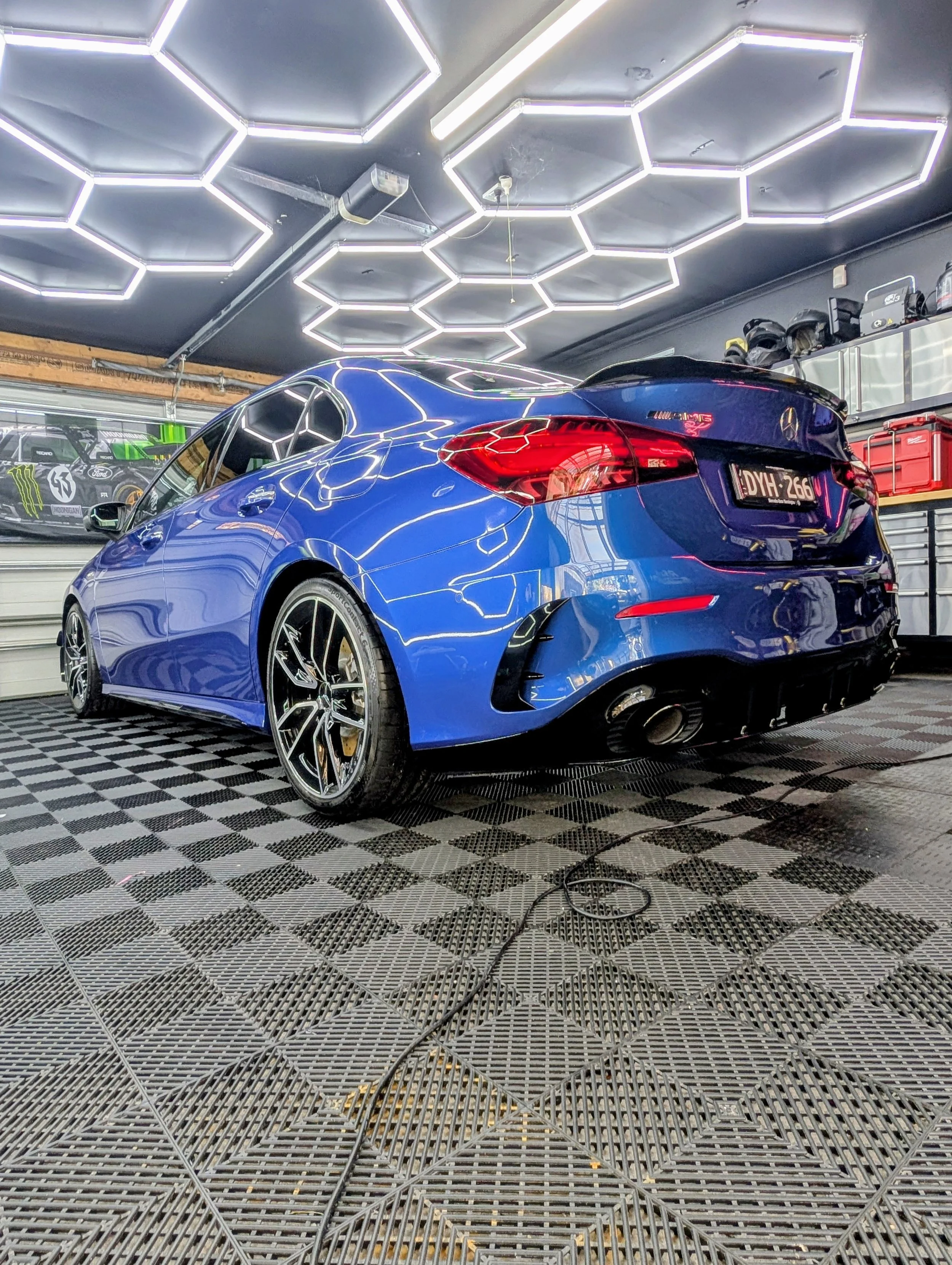 Blue sports car in a garage with modern hexagon-shaped LED ceiling lights and tool storage in the background.