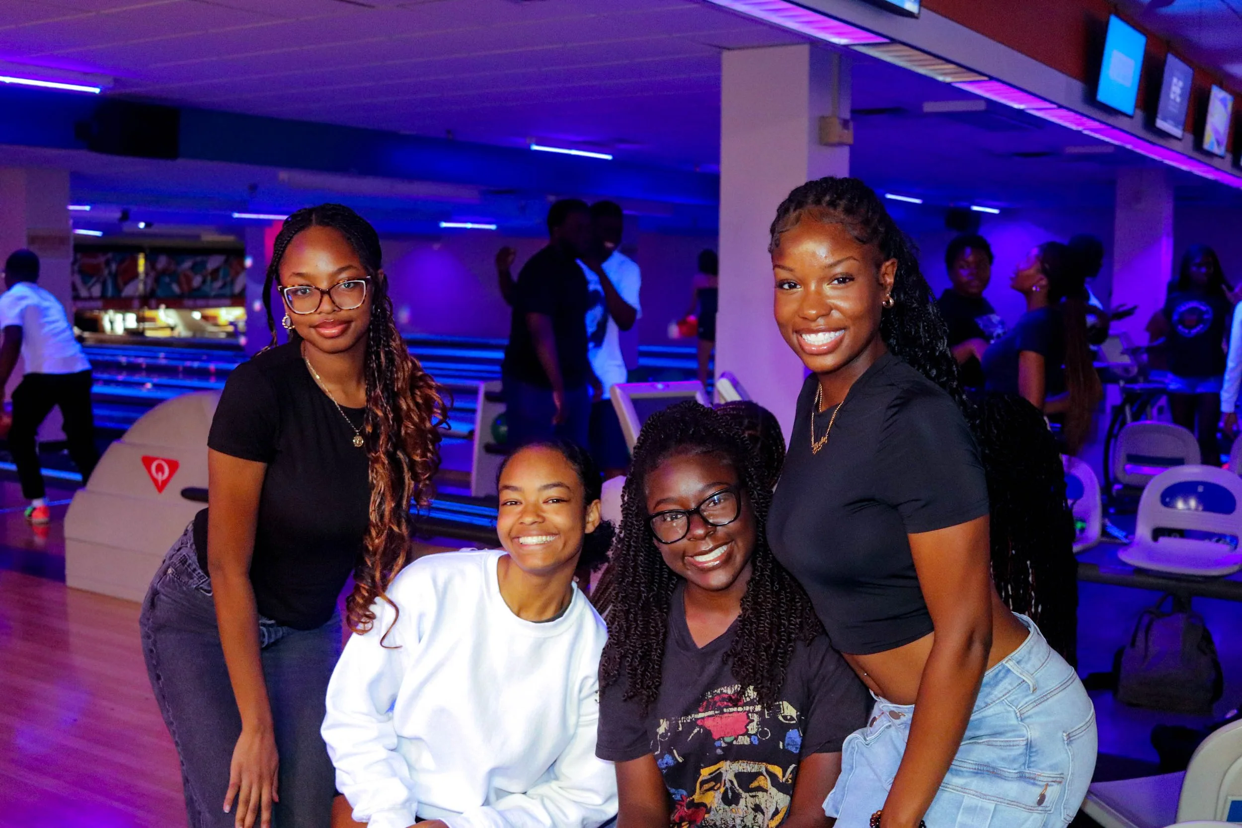 Four friends smiling and posing together in a bowling alley under neon lighting.