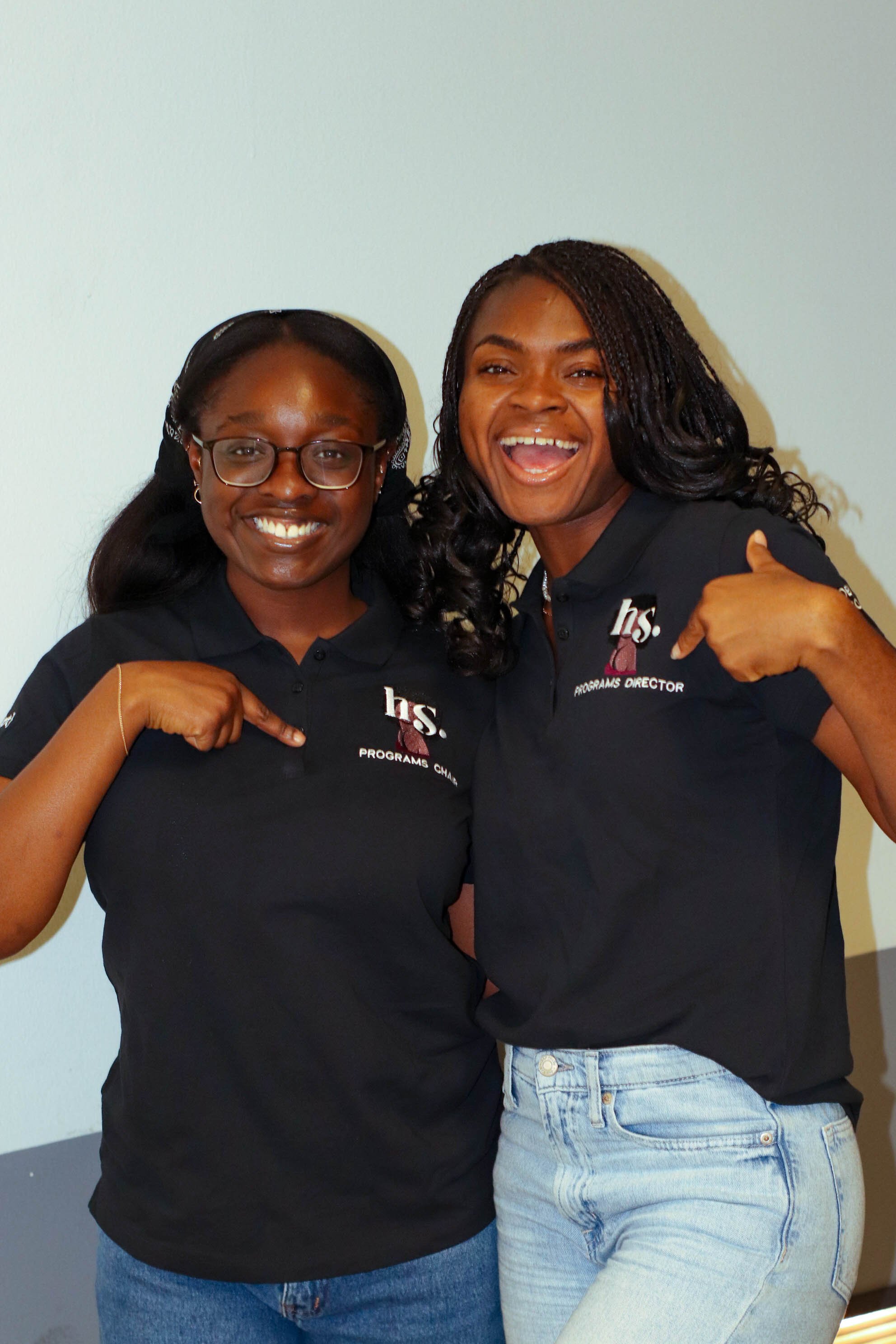 Two women smiling and pointing to their shirts with titles "Programs Chair" and "Programs Director."