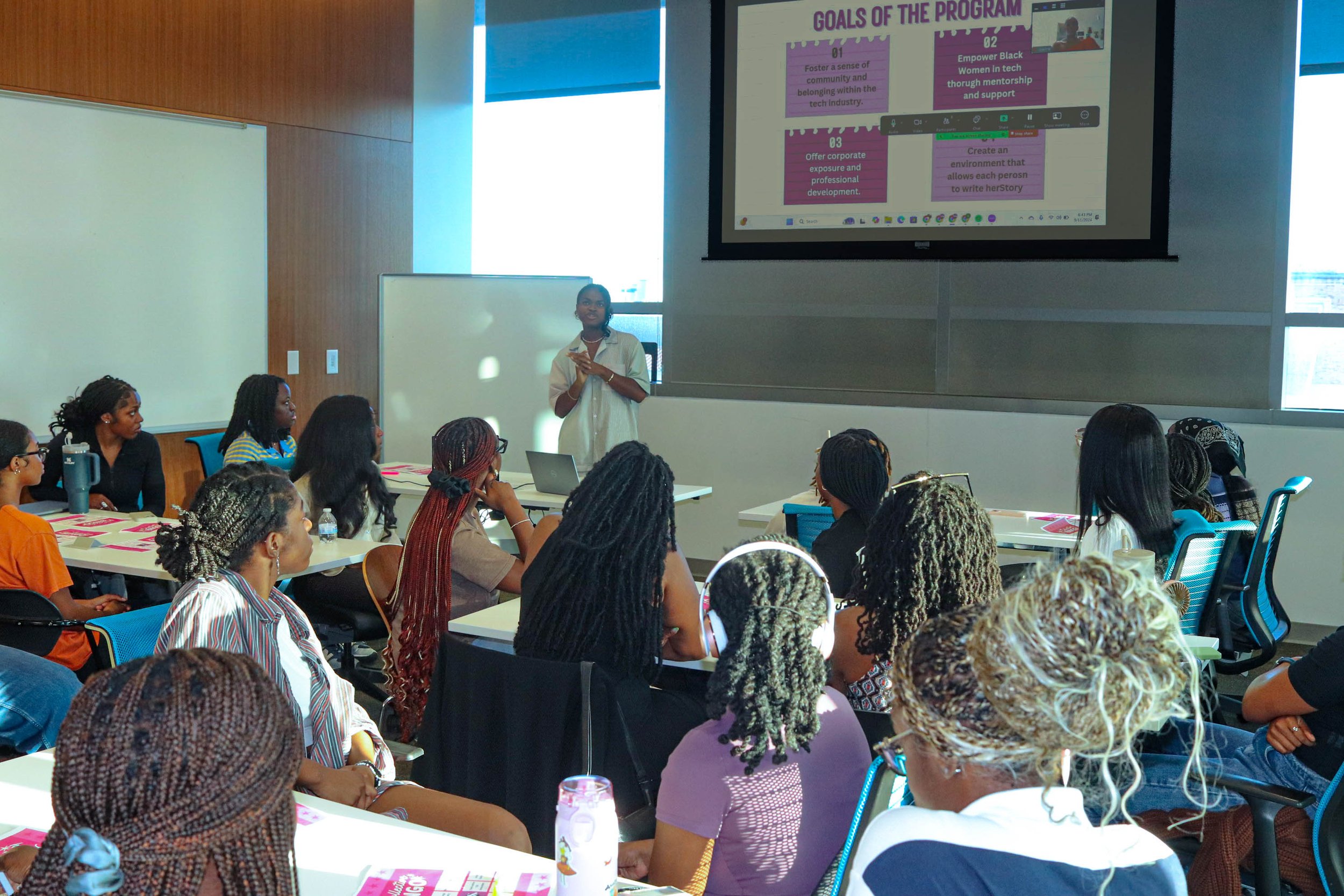 Classroom setting with a presenter addressing attendees, predominantly women, focusing on program goals projected on a screen.
