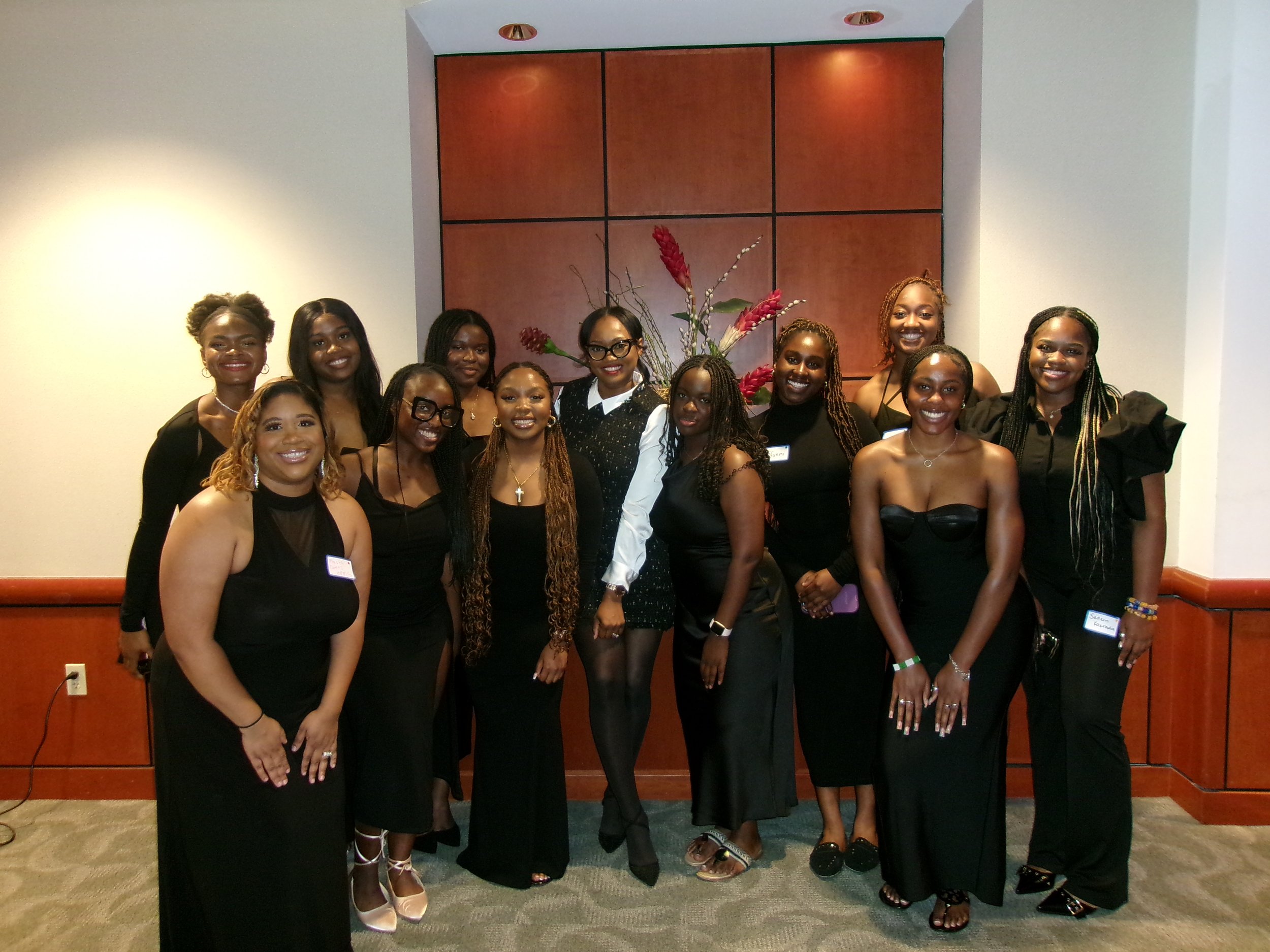 A group of thirteen women posing together indoors, all wearing black dresses, with a decorative arrangement in the background.