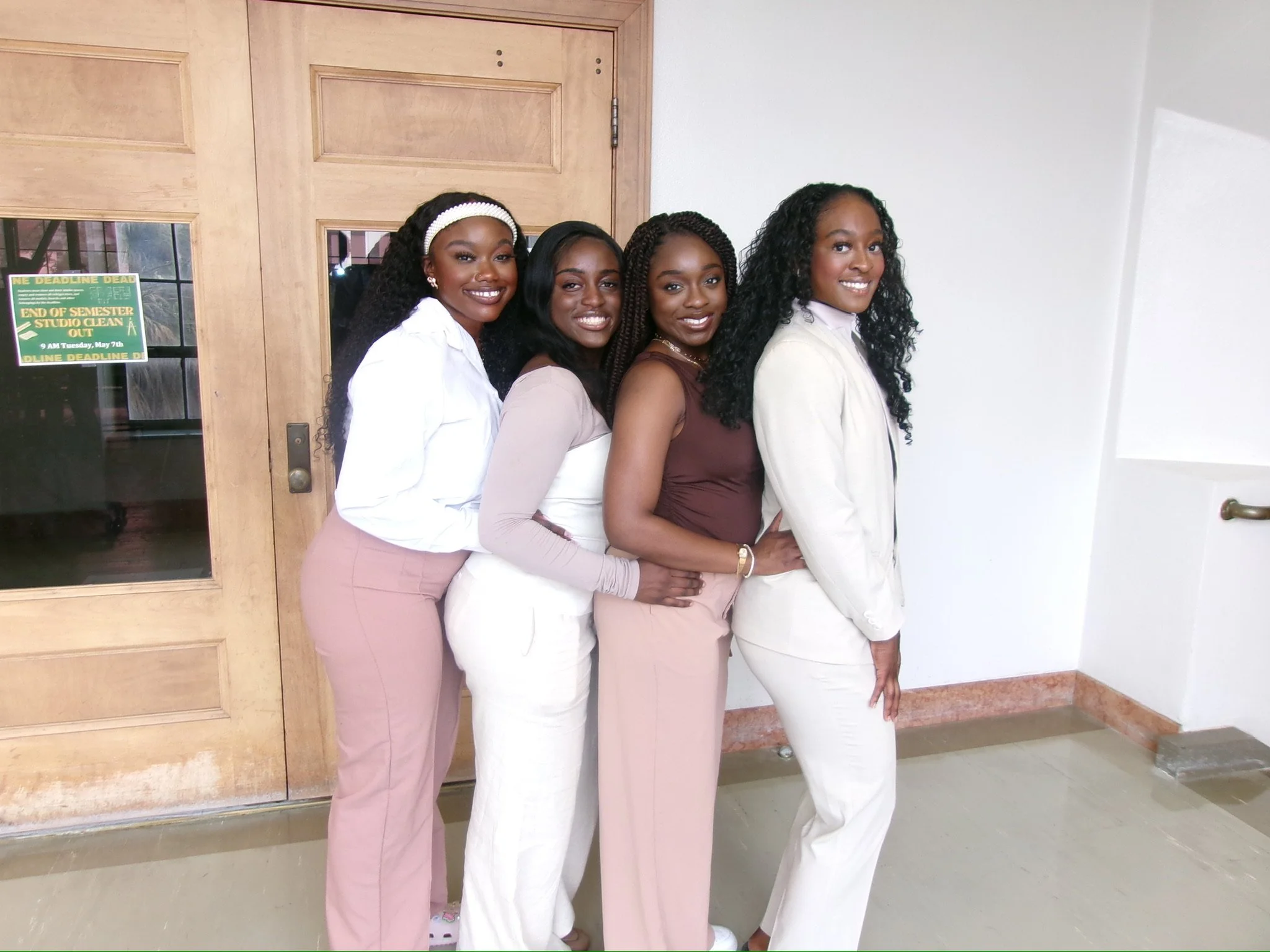 Four women smiling and posing indoors near a wooden door, dressed in business casual attire.